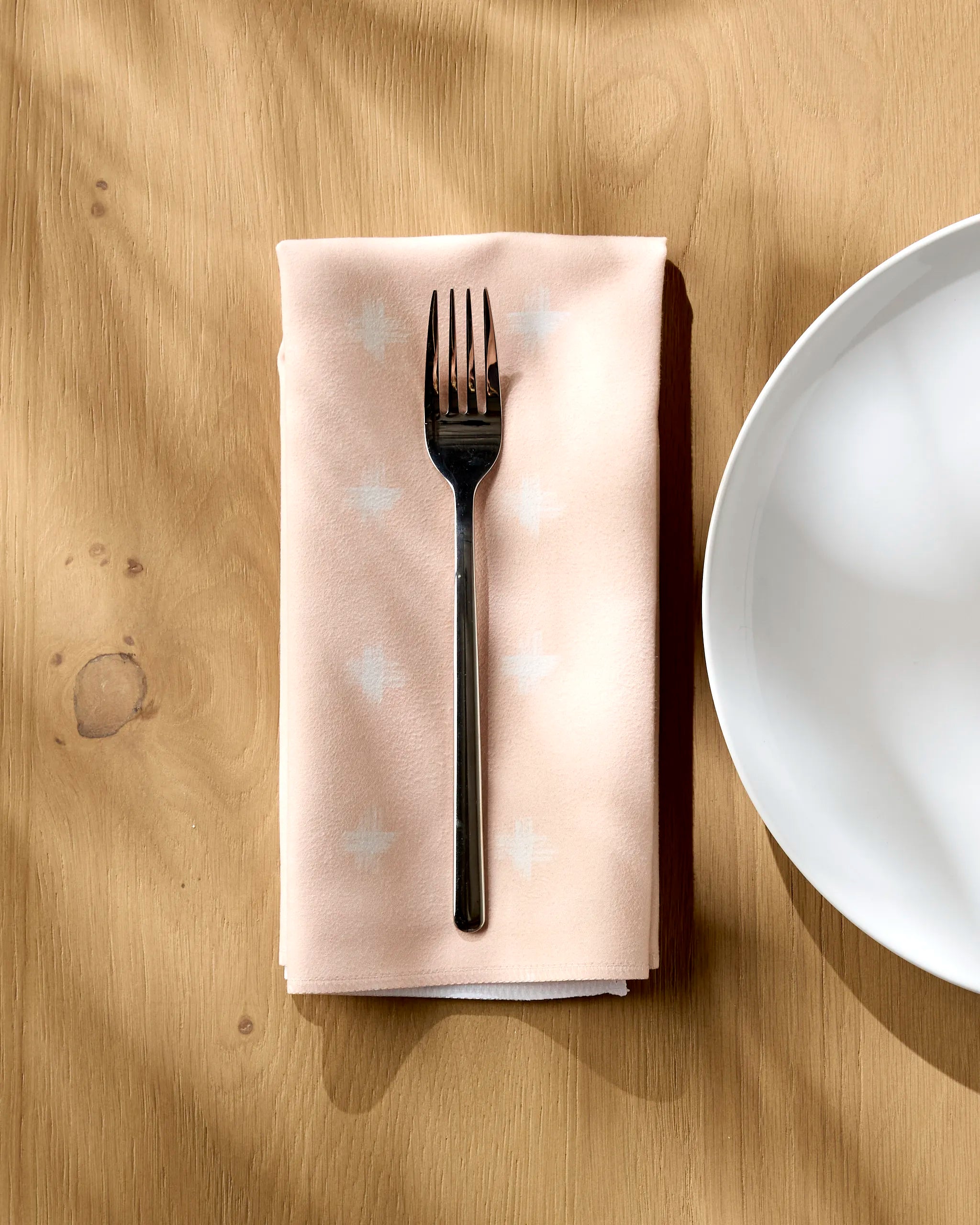 An Aveline dinner napkin by Amy MacCready, featuring subtle white patterns, is folded beneath a silver fork on a wooden table beside a white plate as sunlight casts gentle shadows.