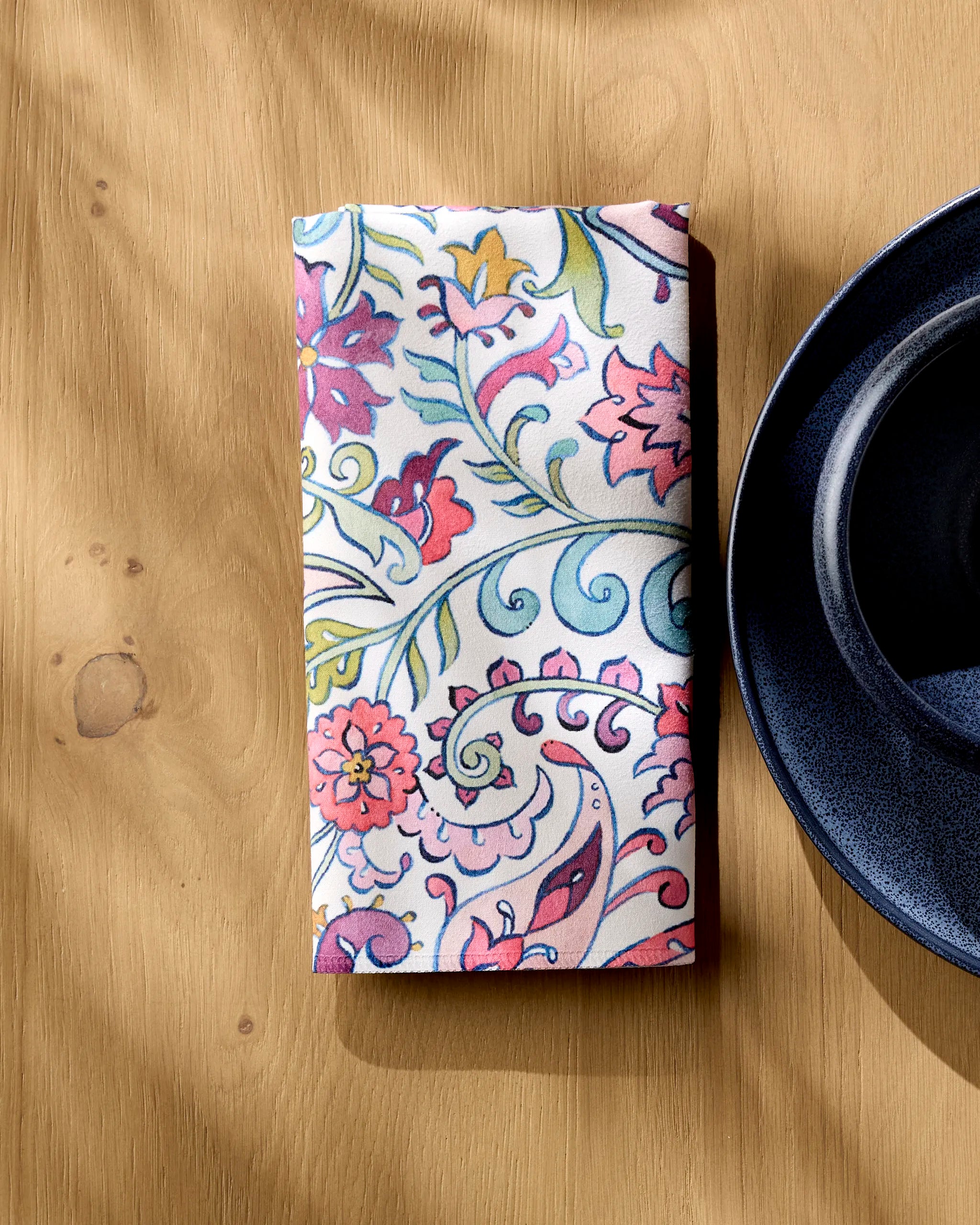 The Bazaar folded washable cloth napkin by Maayan Alper-Swan, featuring a colorful floral pattern, rests on a wooden table beside a dark blue ceramic plate while sunlight casts gentle shadows across the scene.