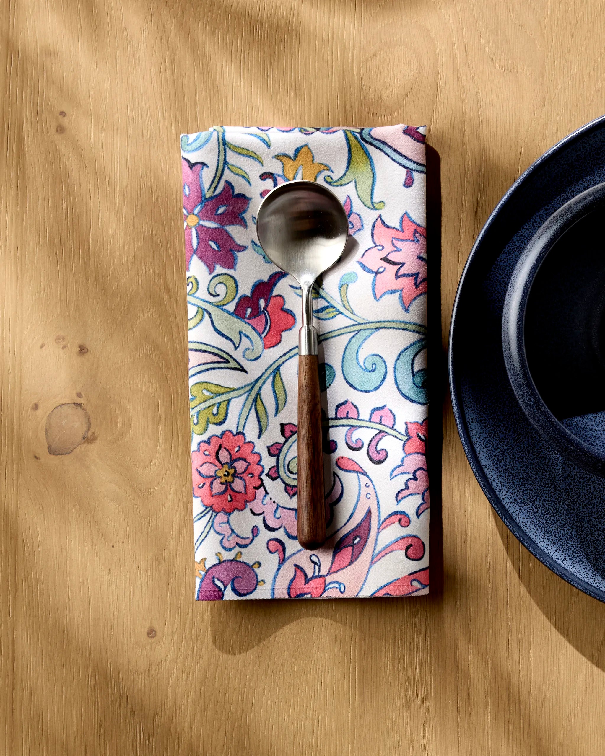 The Bazaar spoon by Maayan Alper-Swan, featuring a dark handle, rests on a colorful floral-patterned washable cloth napkin beside a dark blue plate on a wooden table.
