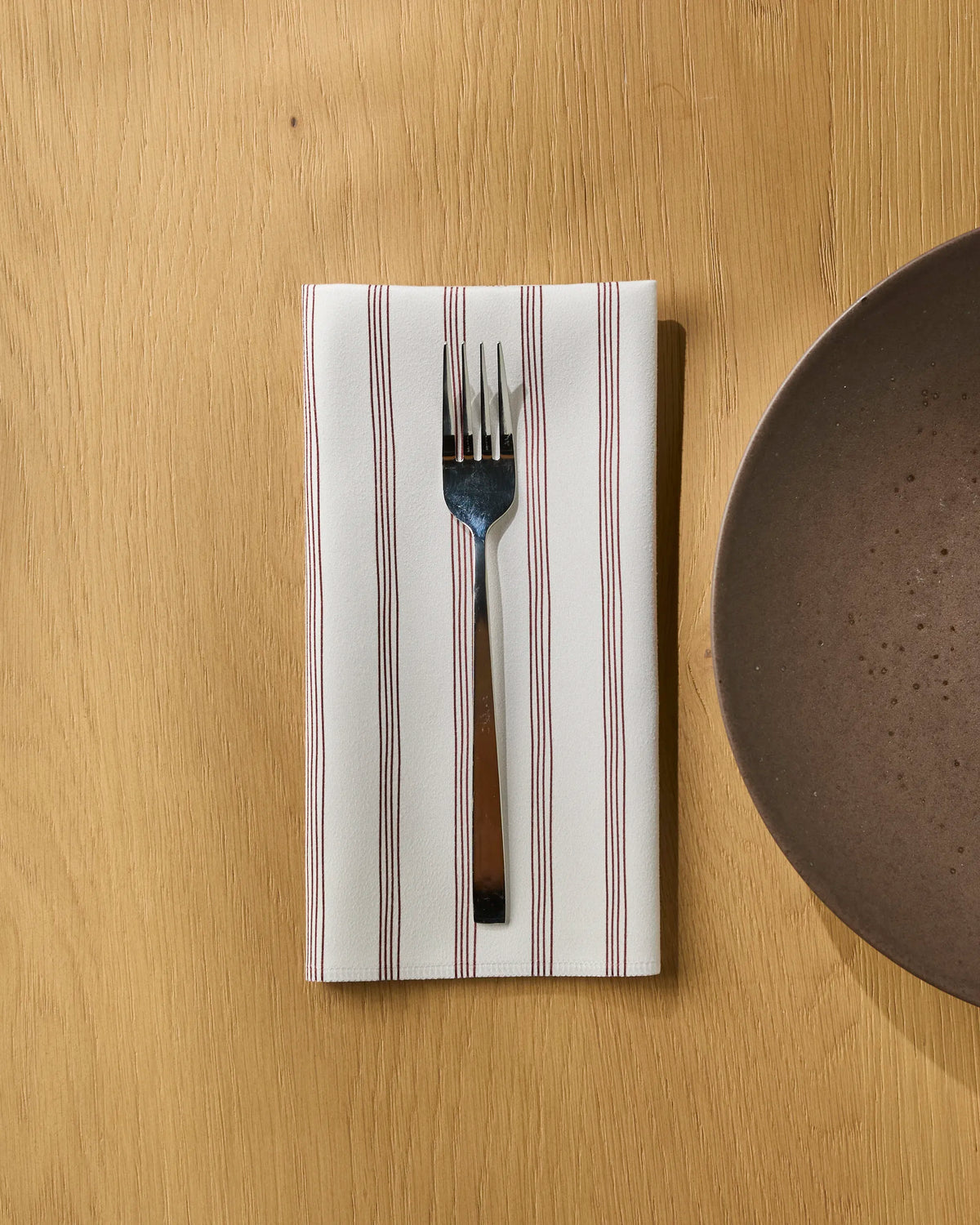 A fork is tucked into a folded Geometry Berry Stripe recycled dinner napkin with red stripes, placed on a light wooden table next to a brown ceramic plate.