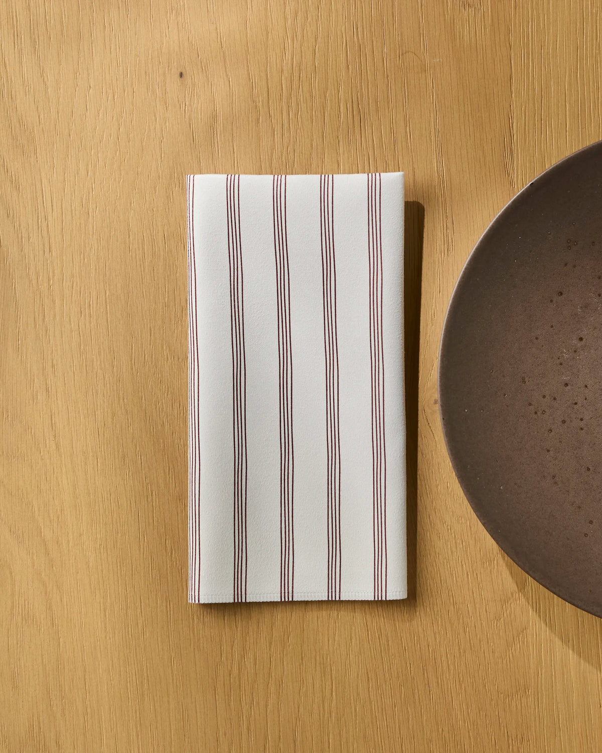 A folded Geometry Berry Stripe napkin, featuring thin red vertical stripes and part of a reusable set, is placed on a light wooden table next to a round brown ceramic plate.