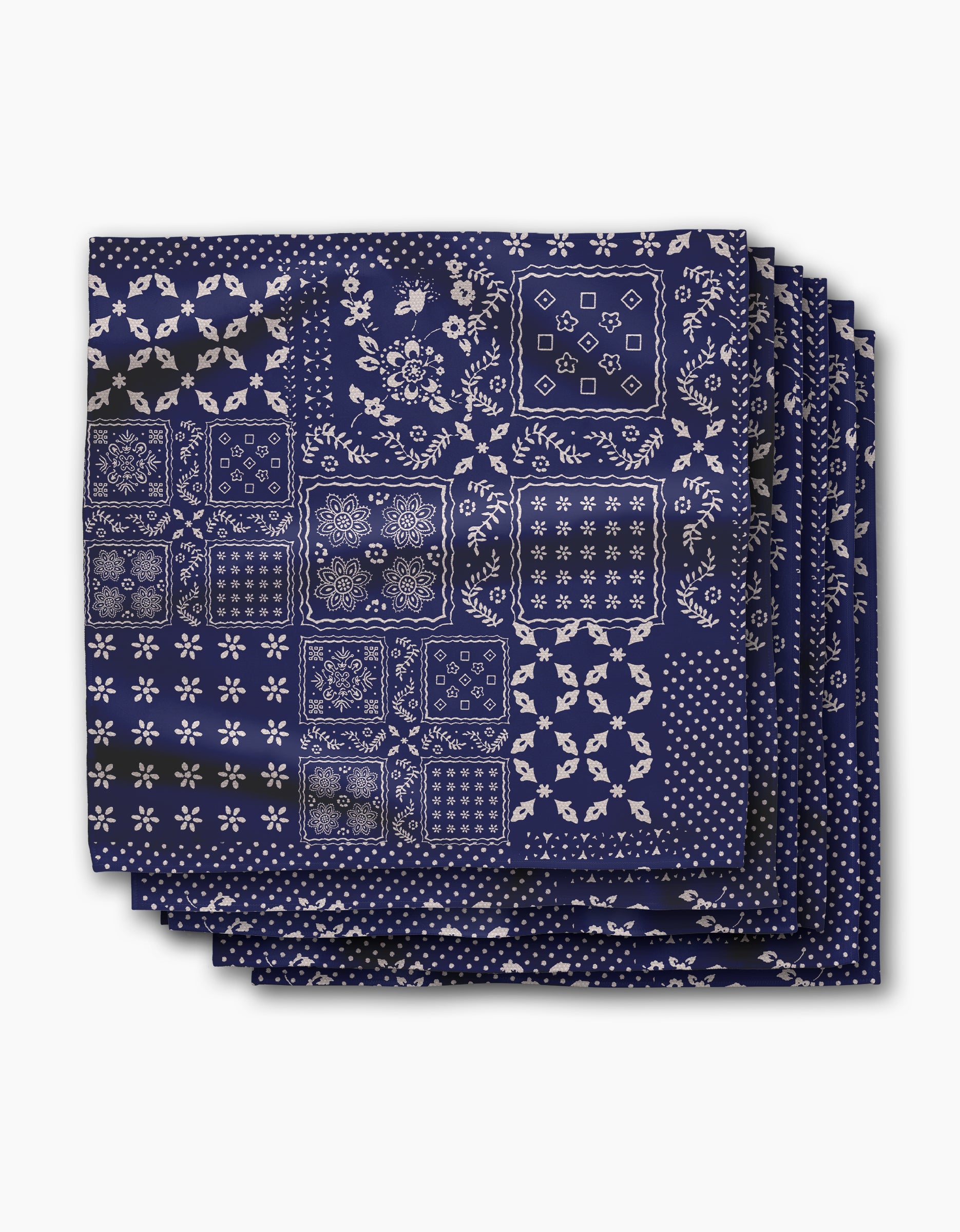 Four stacked Blossoming Bandanna Blue dinner napkins by Geometry, made from recycled geosuede fabric, display assorted white floral and geometric patchwork on navy blue, shown against a white background.
