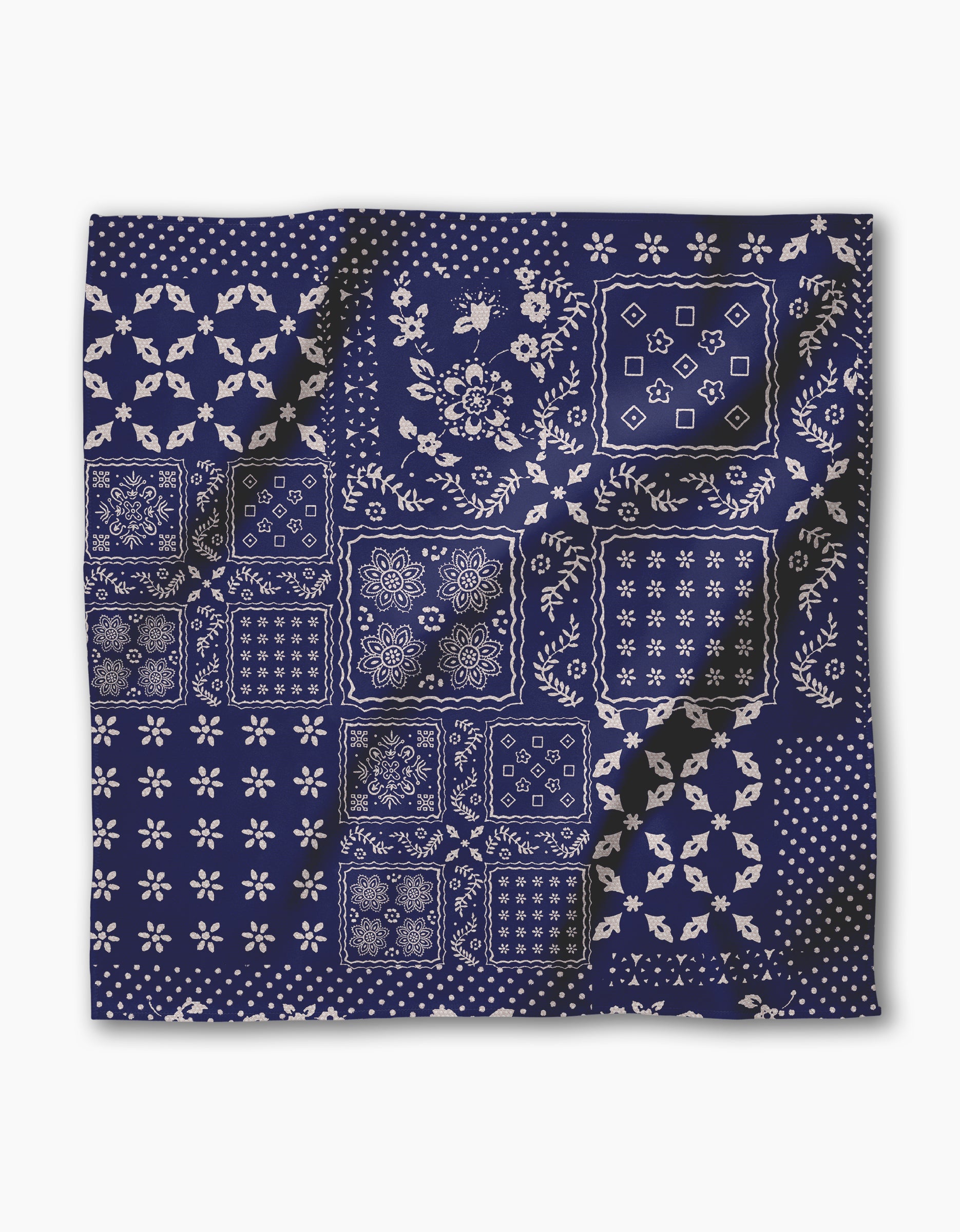 The Blossoming Bandanna Blue by Geometry is a navy blue Geosuede fabric square with white floral, geometric, and polka dot patchwork. Its crumpled texture adds depth, and its an eco-friendly dinner napkin made from recycled materials.