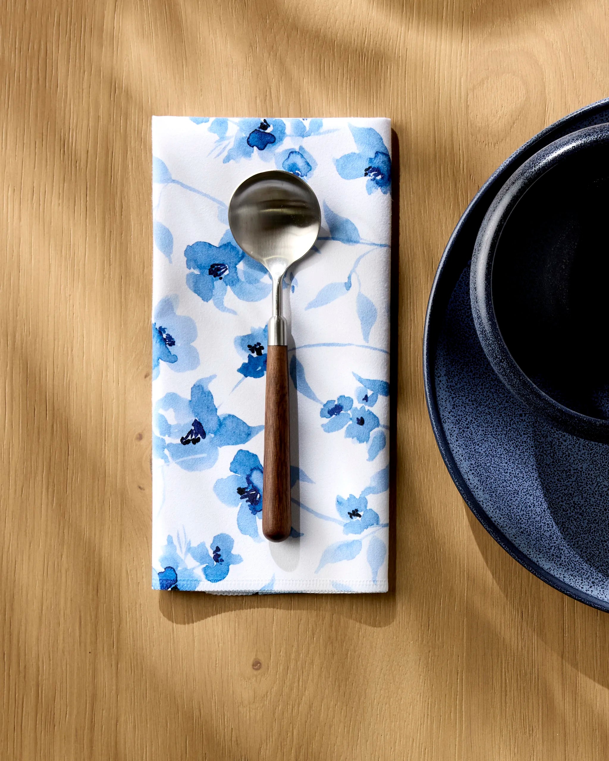 An Aubrey Fairchild "Blue Floral" dinner napkin lies under a spoon with a wooden handle, beside a dark blue bowl and plate on a light wood table.