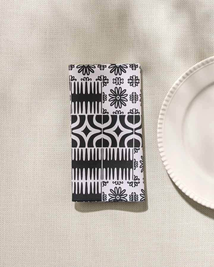 The GEOMETRY Bohemian Mosaic is a folded black and white reusable dinner napkin featuring geometric and floral patterns, shown on a light textured surface beside a scalloped white plate.