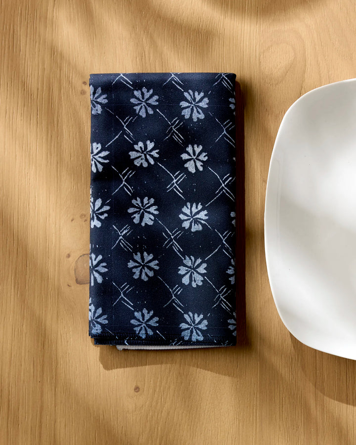The Geometry Bonnie Indigo napkin, a folded navy blue reusable with white floral and grid patterns, rests on a light wooden table beside a white plate. Sunlight highlights this elegant recycled fabric accessory.