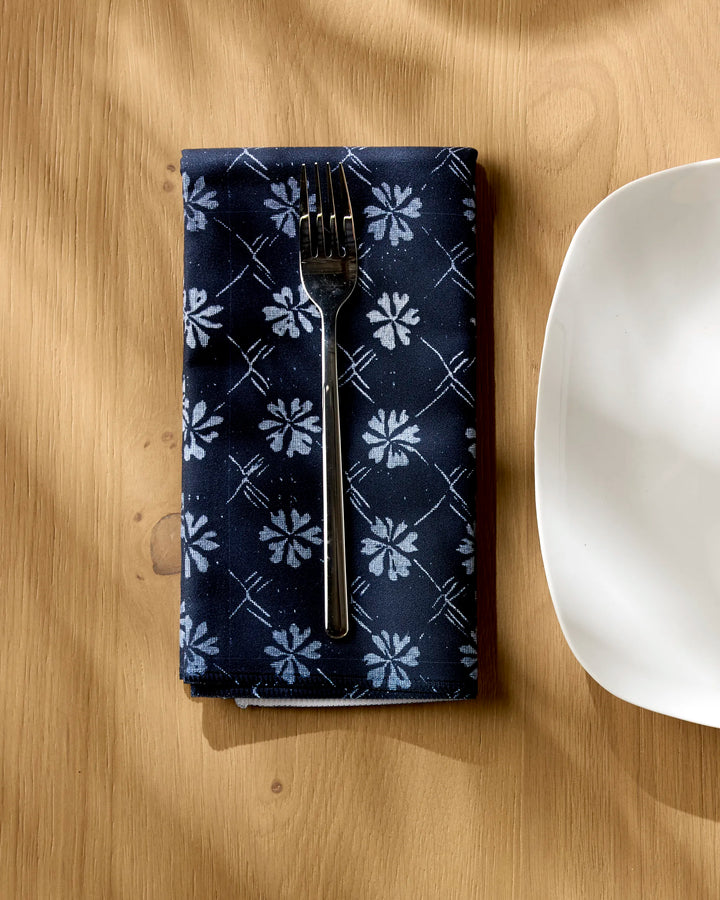 A fork rests on the Bonnie Indigo reusable napkin by Geometry, featuring a navy blue floral pattern, beside a white square plate on a light wooden table with shadows across the surface.