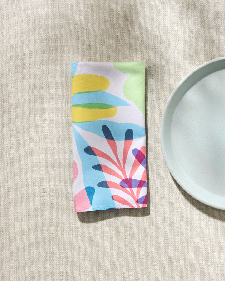 The GEOMETRY "Botanical Burst" reusable napkin, featuring colorful abstract plant designs, rests next to a light blue ceramic plate on a textured beige surface, softly lit by natural light.