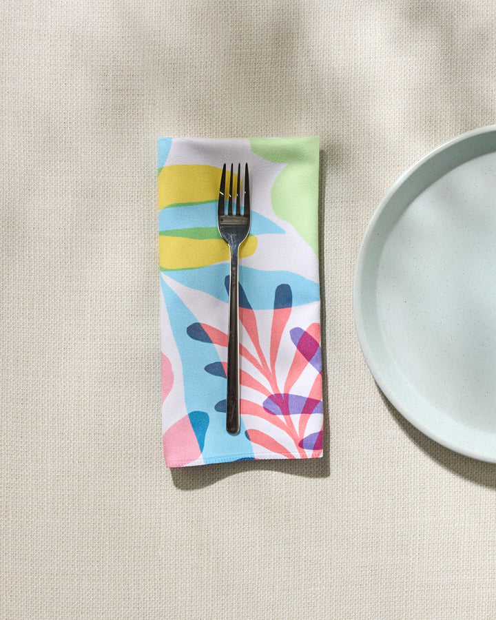 A metallic fork rests on GEOMETRY's Botanical Burst dinner napkin, featuring colorful abstract shapes, beside a plain white plate on a textured, light-toned surface.