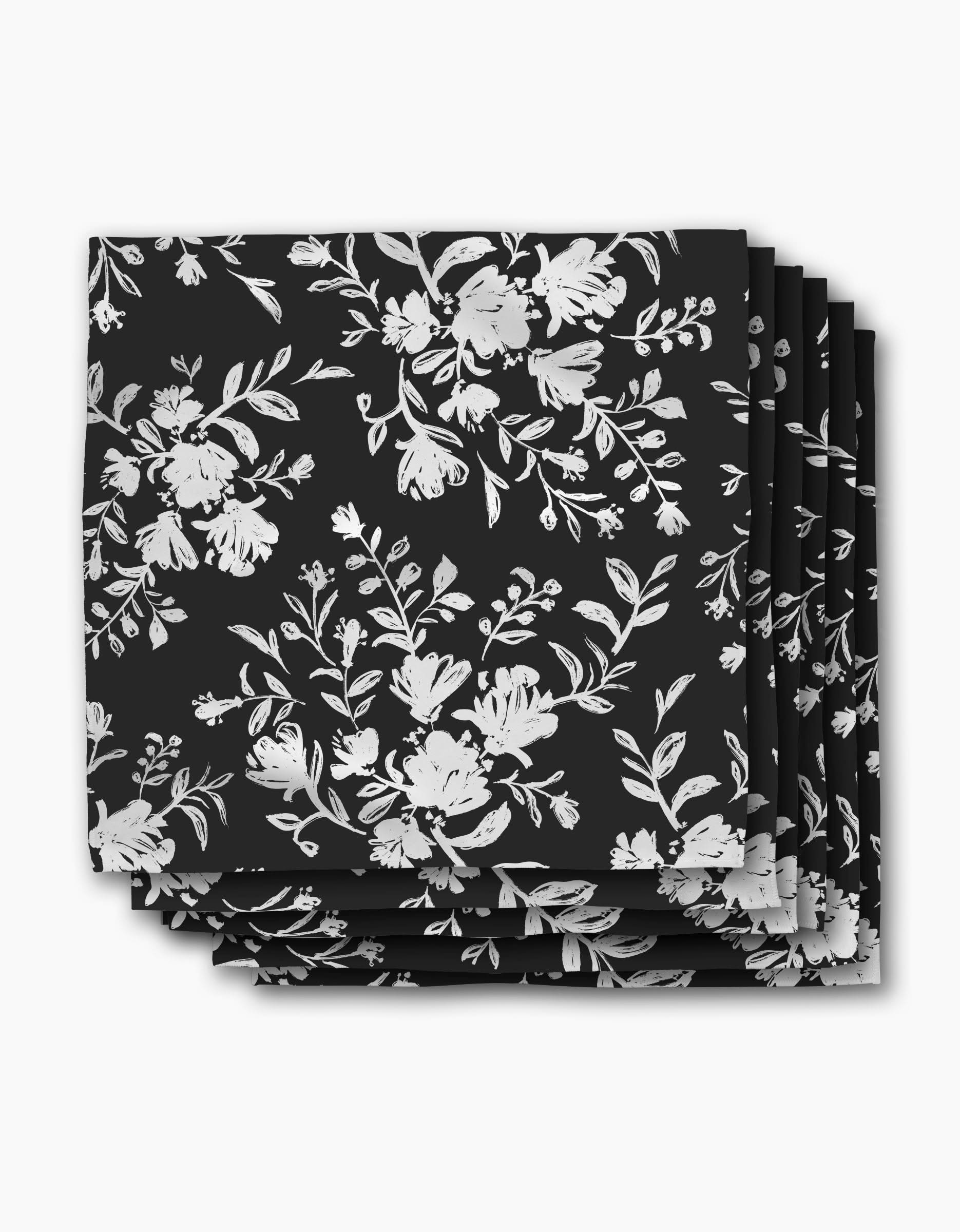 A stack of Danelys Sidron Botanical Silhouettes Charcoal dinner napkins, made from soft Geosuede fabric and featuring a white floral and leaf design, neatly arranged against a white background.