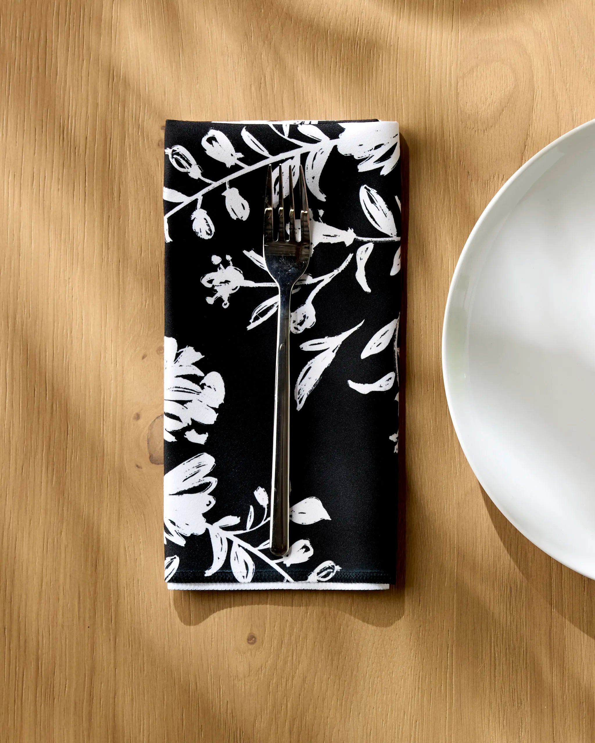 A silver fork rests on a Danelys Sidron Botanical Silhouettes Charcoal black recycled dinner napkin with white floral patterns, set on a wooden table next to a white plate, as sunlight casts soft shadows over the absorbent napkin.