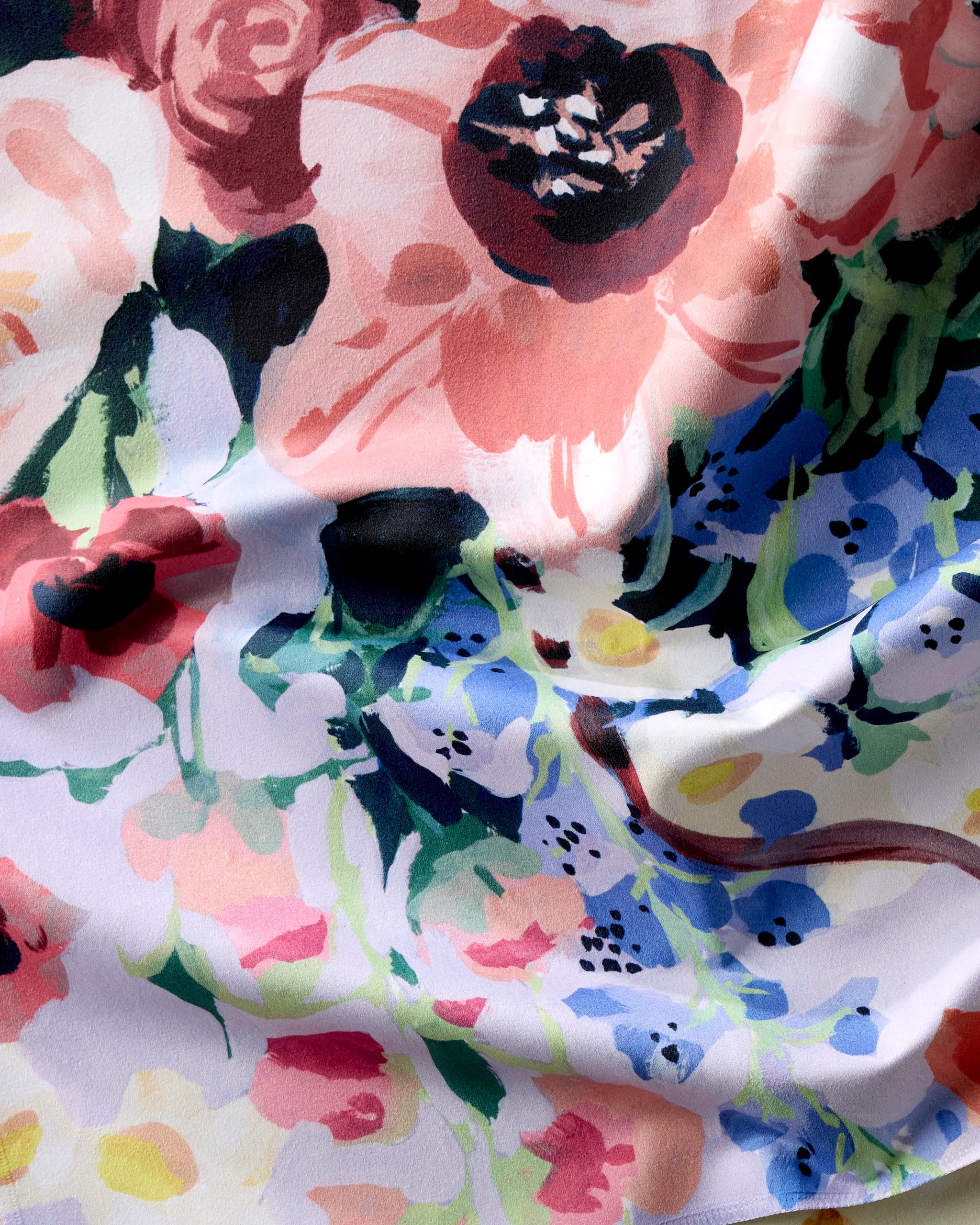 A close-up of Ania Zwara Art's Bouquet reusable cloth napkin shows its soft white fabric, gently wrinkled, with a watercolor-style floral pattern in pink, red, blue, yellow, and green.