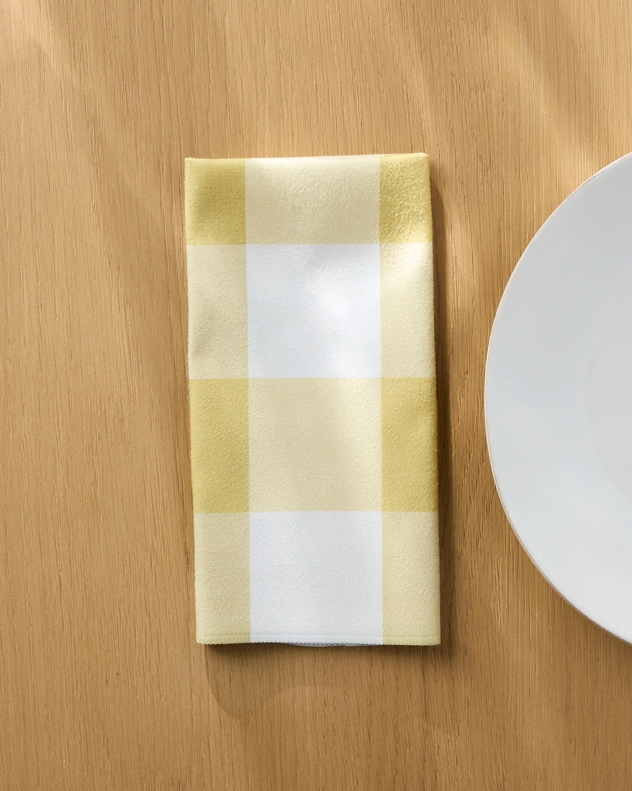 The Bright Gingham reusable cloth napkin by Jasmine Blooms Designs, featuring yellow and white checks, is folded on a light wooden table beside a plain white plate.