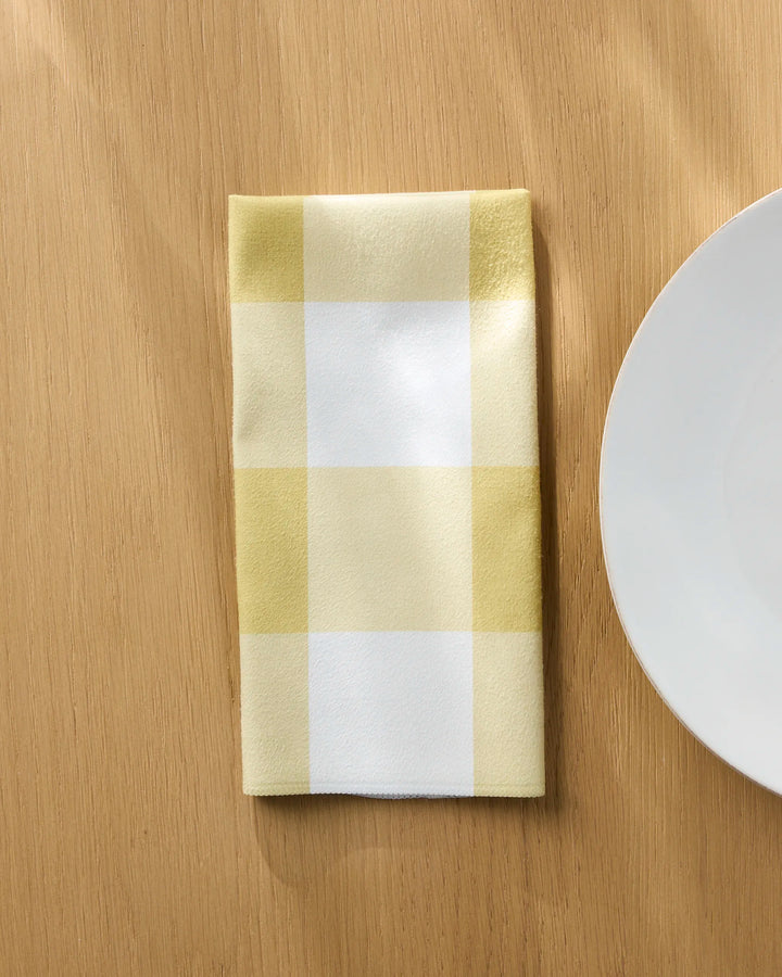 The Bright Gingham reusable cloth napkin by Jasmine Blooms Designs, featuring yellow and white checks, is folded on a light wooden table beside a plain white plate.