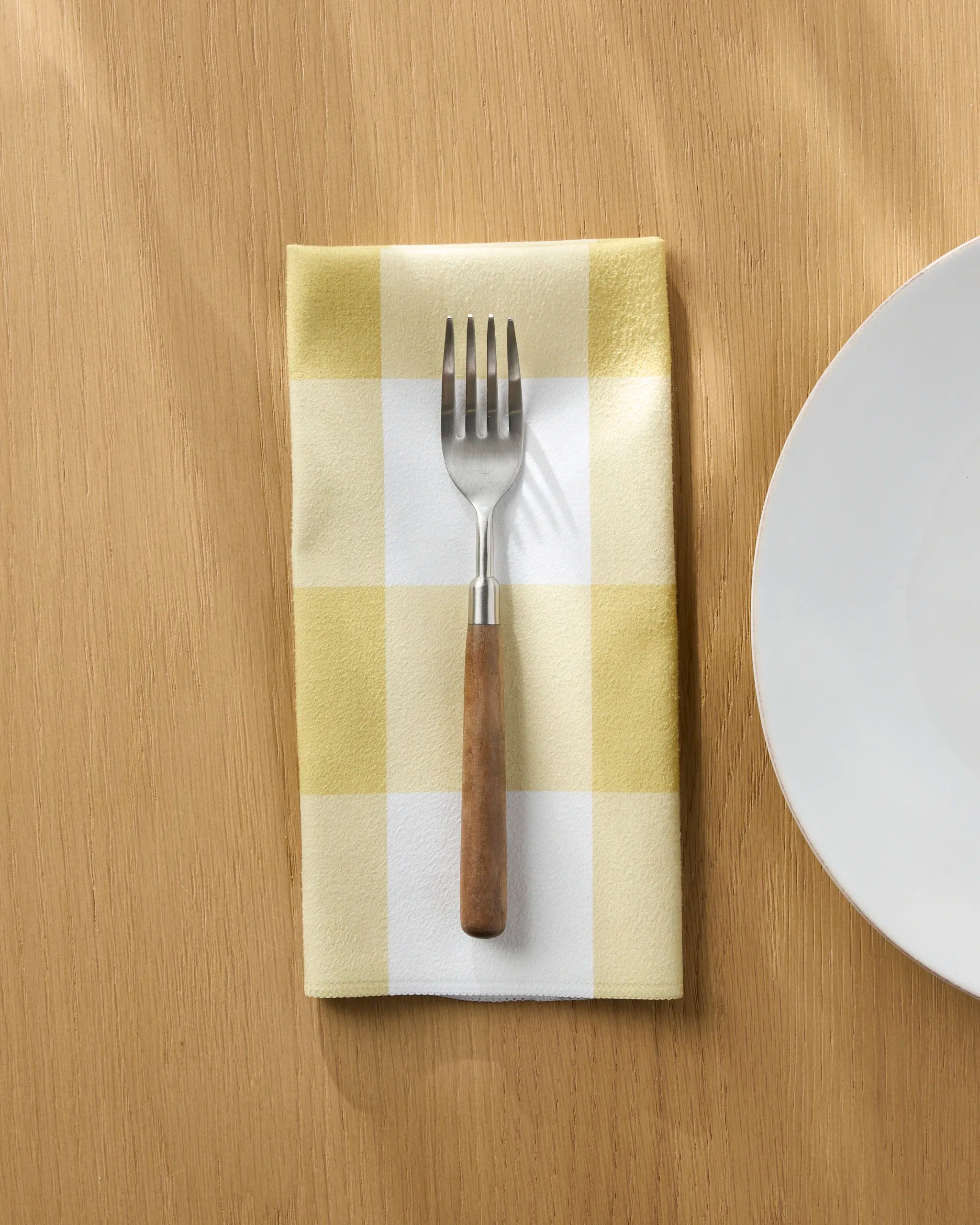 A Jasmine Blooms Designs Bright Gingham yellow and white checkered reusable cloth napkin rests beneath a fork with a wooden handle on a light wooden table beside a white plate.