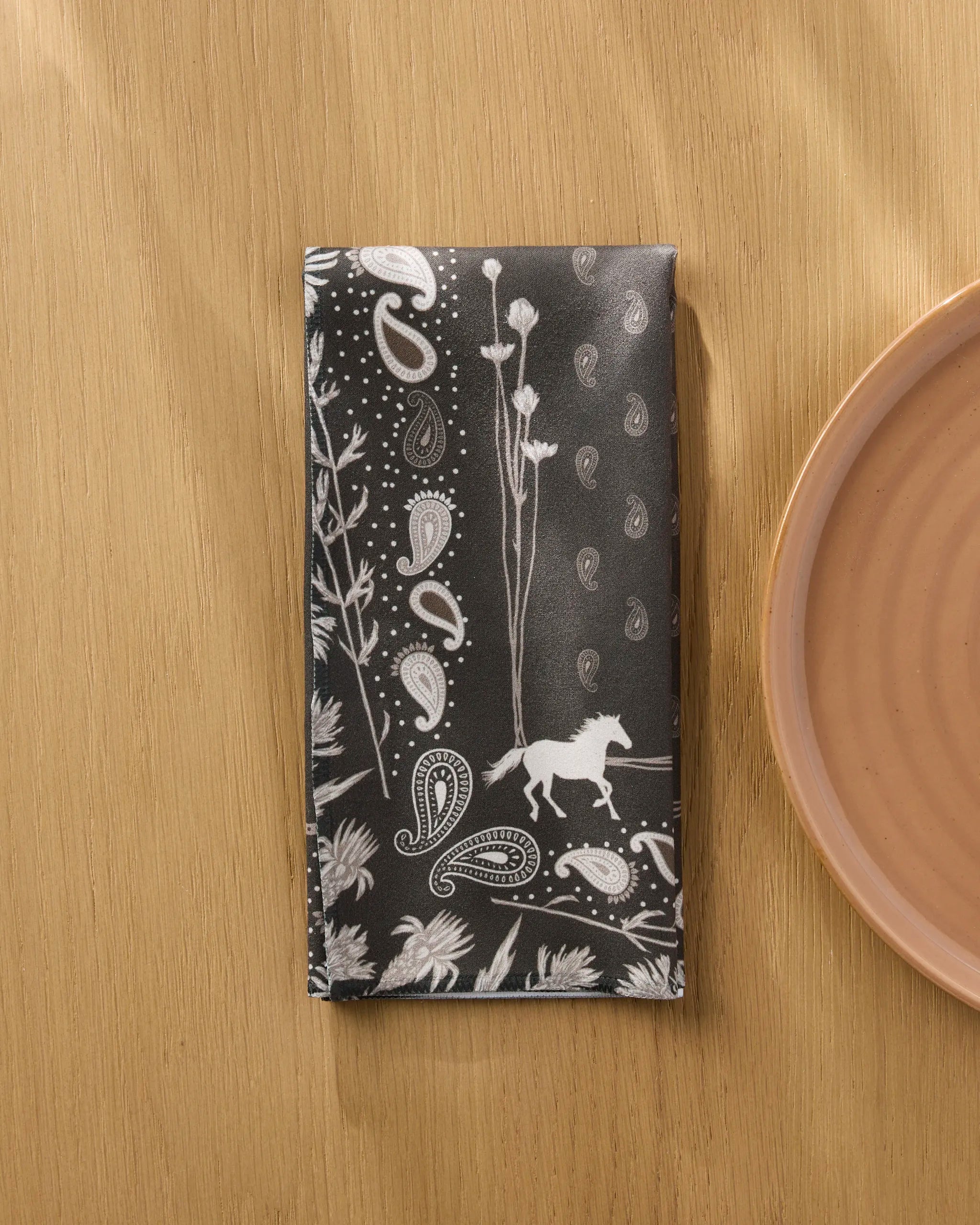 The Geometry Bronco Espresso napkin, featuring black Geosuede fabric with white paisley, plants, and a horse design, is displayed on a wooden table beside a pink ceramic plate.