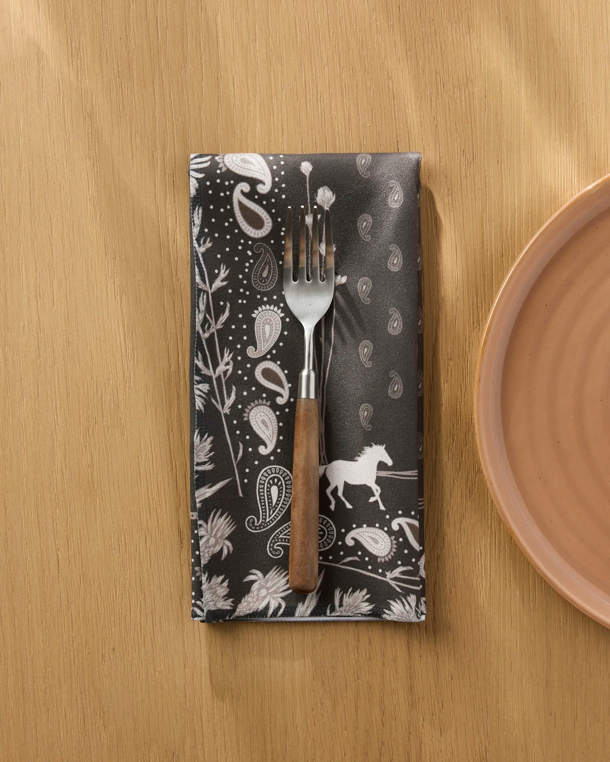 A fork with a wooden handle rests on Geometry's Bronco Espresso recycled dinner napkin, featuring paisley and horse motifs, beside a peach plate on a wooden table.