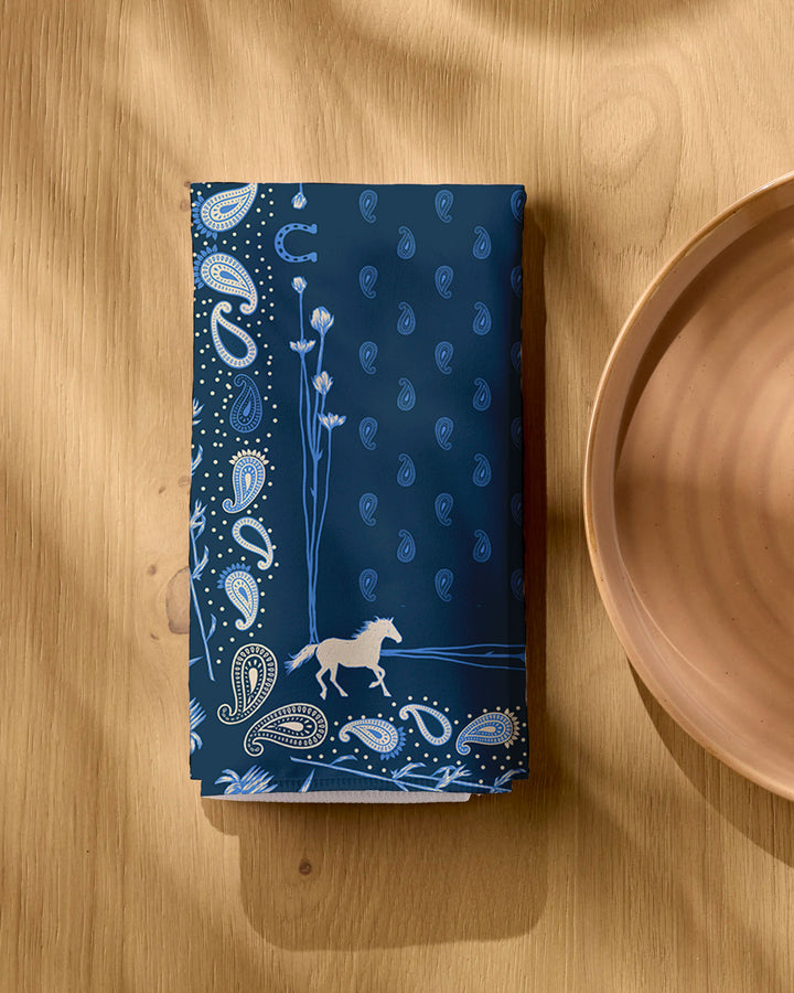A folded Bronco Indigo napkin from Geometry, featuring a light blue paisley pattern and white horse design, rests on a wooden table near a tan ceramic plate, with shadow patterns visible across the surface.