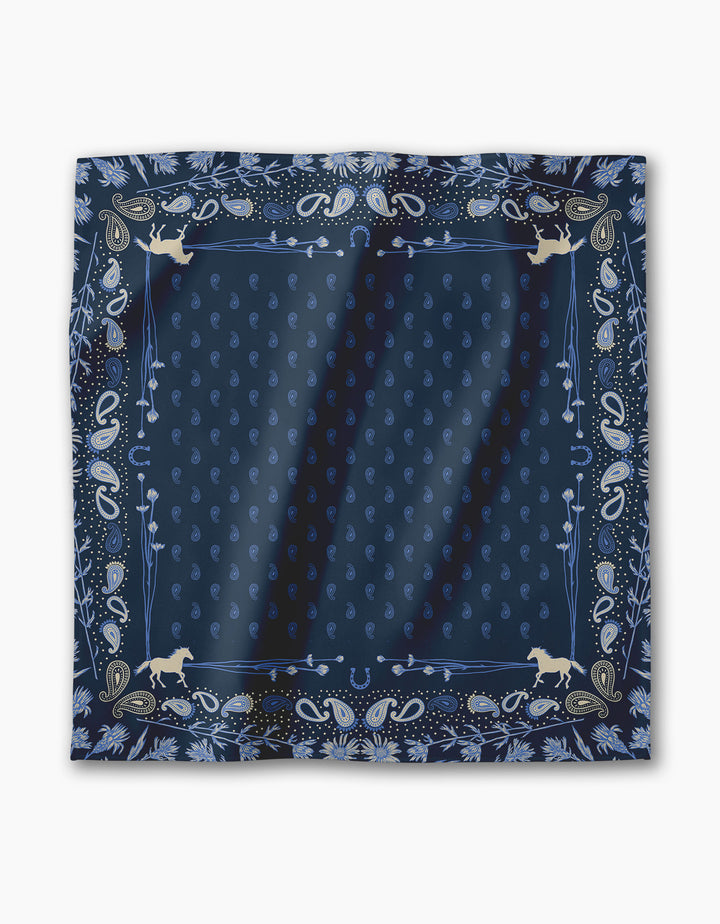The Bronco Indigo by Geometry is a navy blue bandana featuring paisley and floral patterns with four tan horse silhouettes in each corner, bordered by intricate designs—perfect as a unique, reusable dinner napkin for gatherings.