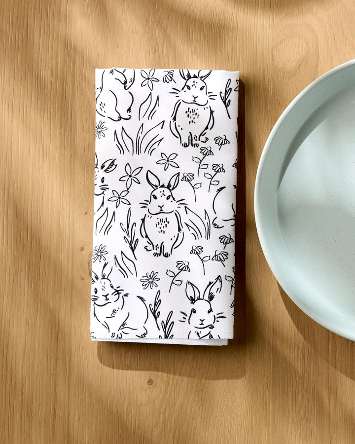 A "Bunny Rabbit Garden" recycled dinner napkin by Charlie Rowan Designs, featuring black bunny and flower sketches, is folded on a light wooden table next to a light blue plate.