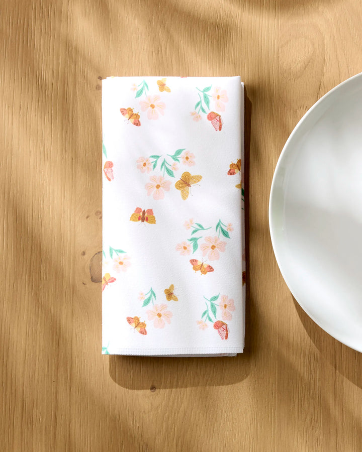The "Butterfly Garden" cloth napkin by Danelys Sidron, featuring a floral and butterfly print, is neatly folded on a wooden table beside a white plate, with sunlight casting gentle shadows over the scene.