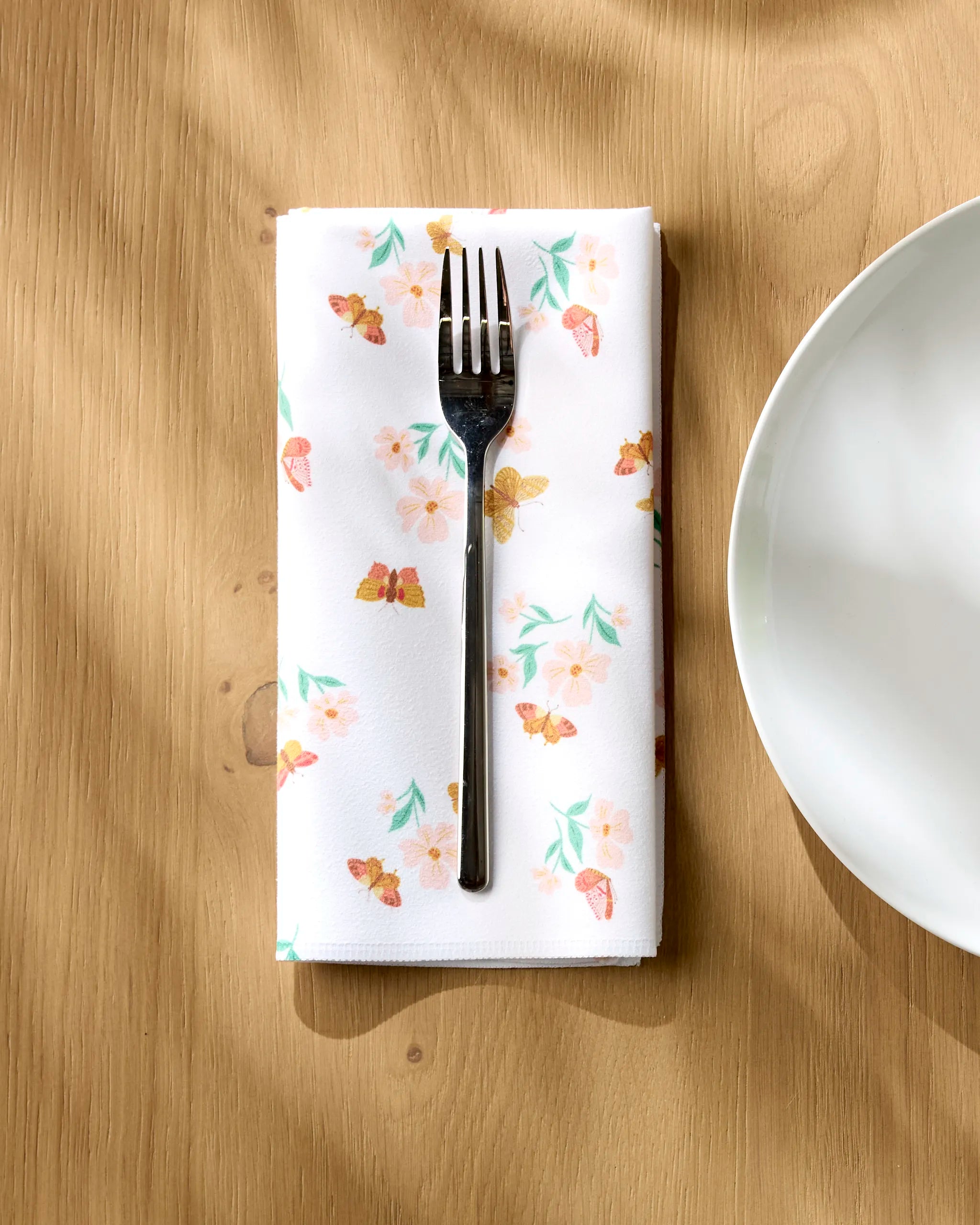 A silver fork rests on an absorbent Butterfly Garden dinner napkin by Danelys Sidron, featuring a floral and butterfly design, placed beside a white plate on a light wooden table as sunlight casts soft shadows.
