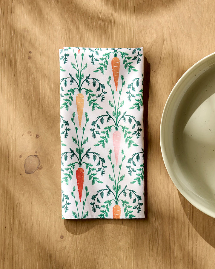 A folded Carrot Damask dinner napkin by Sarah Daugherty, featuring a carrot and green leaf pattern on soft Geosuede, rests on a light wooden table beside a beige ceramic bowl as sunlight casts gentle shadows.