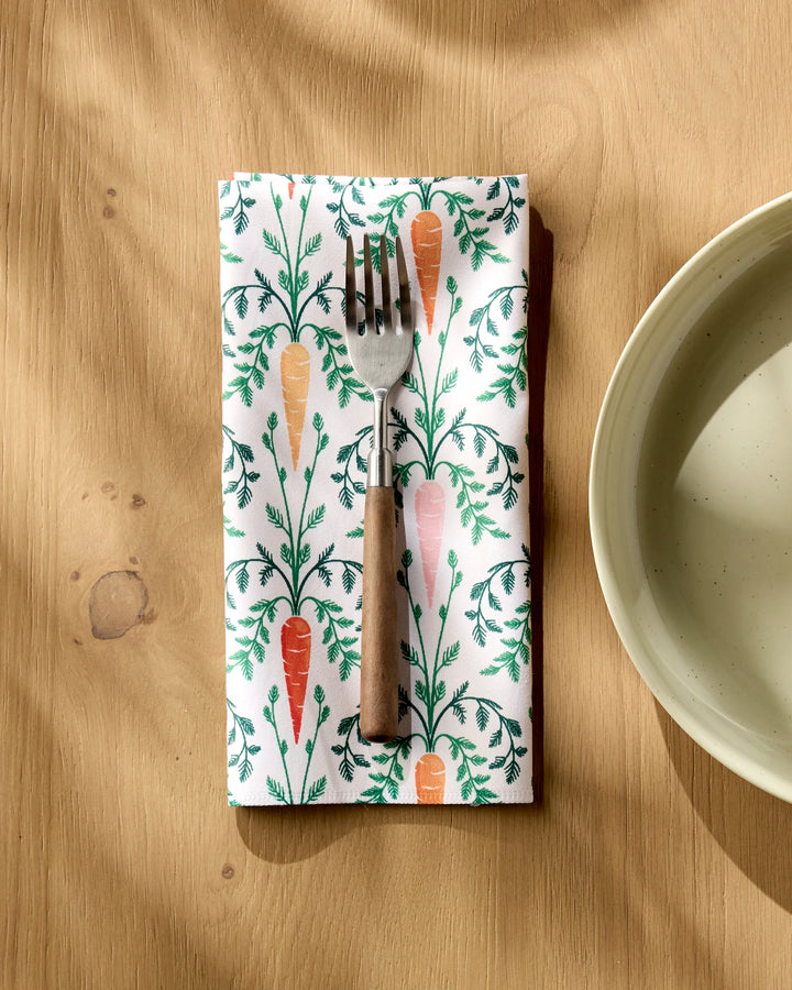 A Sarah Daugherty Carrot Damask recycled napkin with carrot and leaf designs holds a wooden-handled fork beside a ceramic bowl on a wooden table.