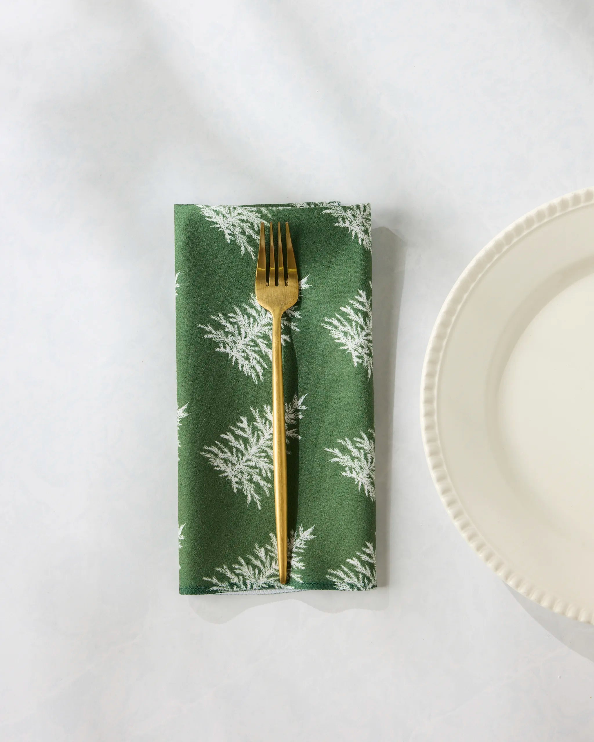 A gold fork rests on the Geometry "Cedar Green" reusable napkin, featuring a white leaf pattern, next to a scalloped-edge white plate on a light surface.
