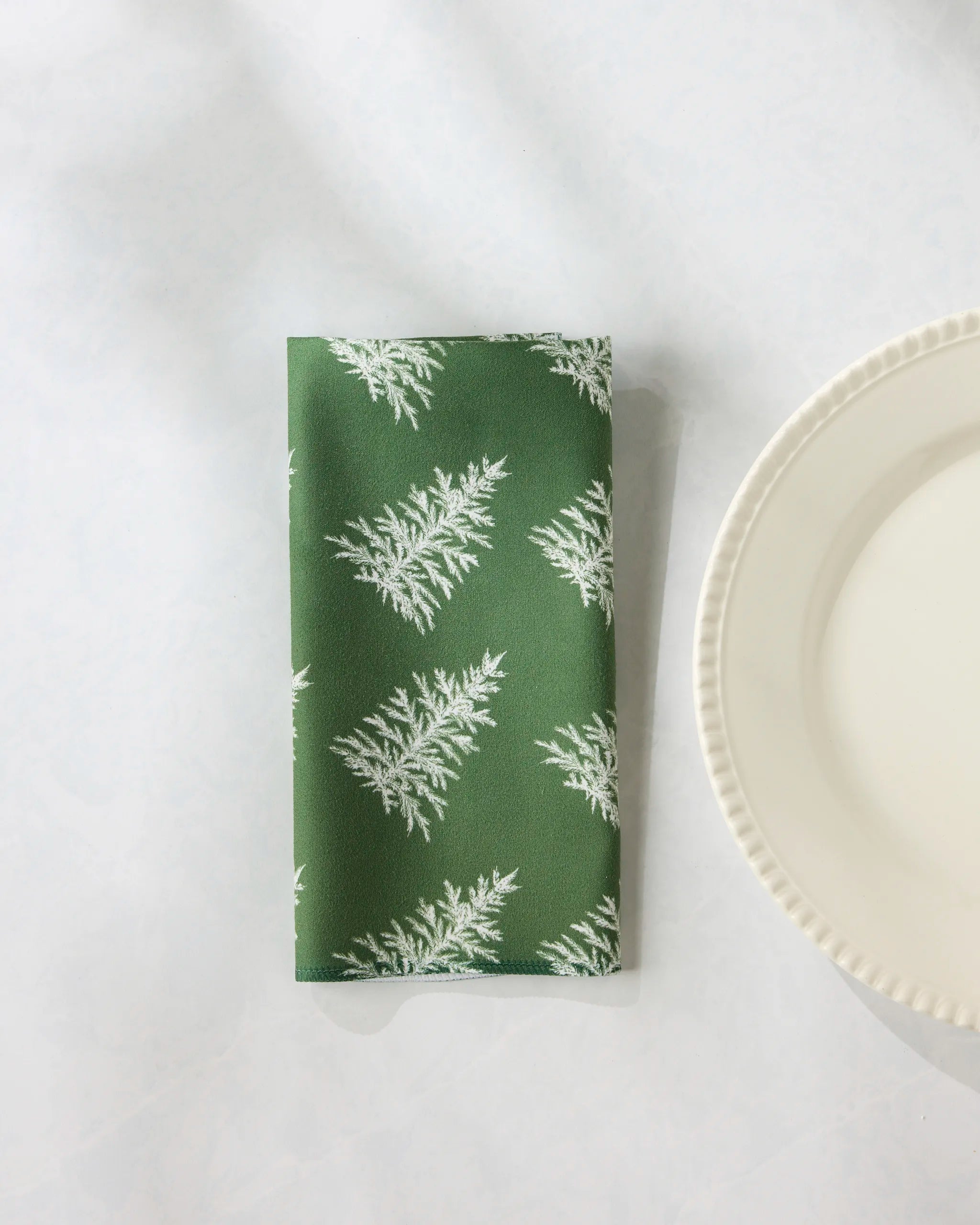 A Cedar Green reusable table napkin by Geometry, featuring a white fern pattern, is folded next to a white plate with scalloped edges on a light surface.
