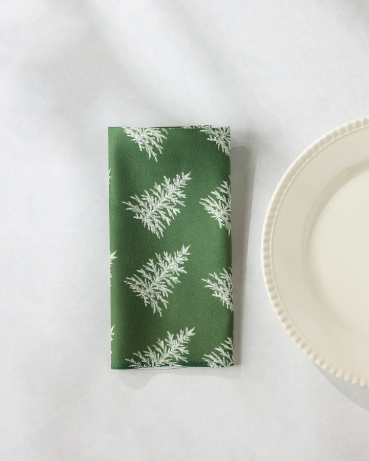 A Cedar Green reusable table napkin by Geometry, featuring a white fern pattern, is folded next to a white plate with scalloped edges on a light surface.