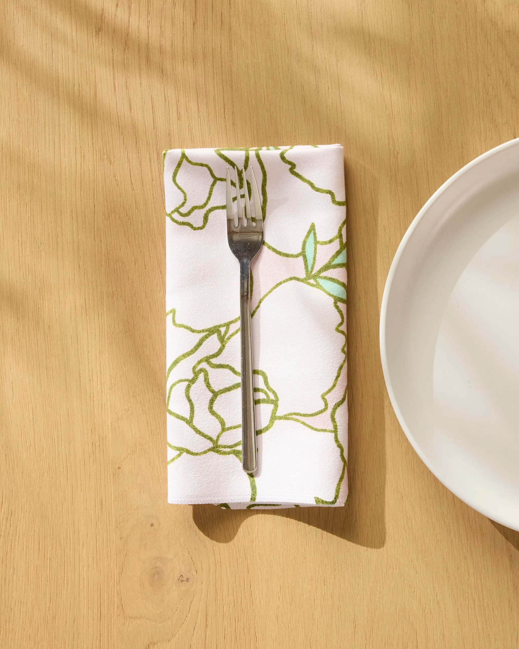 The Celecia reusable napkin by Amy MacCready, with its green floral pattern, is folded under a fork beside a white plate on a pale wood table, as sunlight casts gentle shadows across the scene.