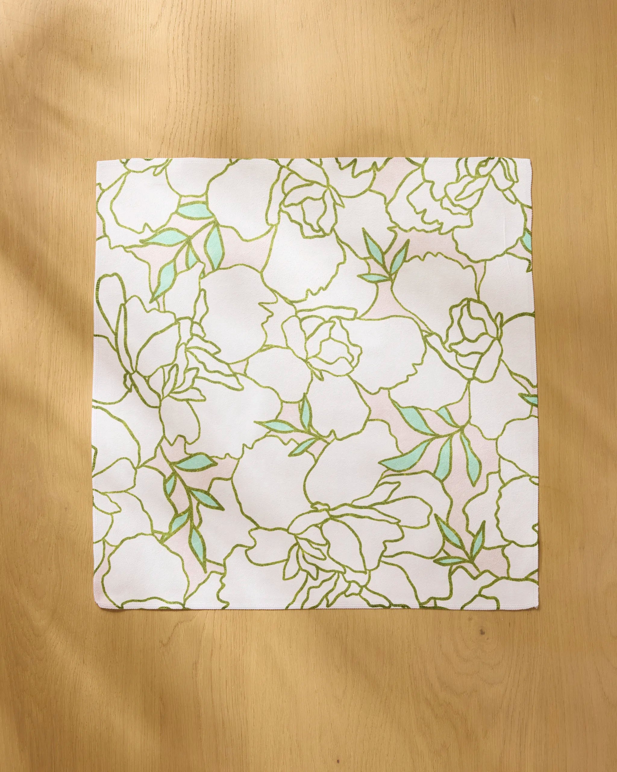 The Celecia napkin by Amy MacCready, a square white reusable piece with a green and gold floral outline pattern, is displayed flat on a light wooden surface.