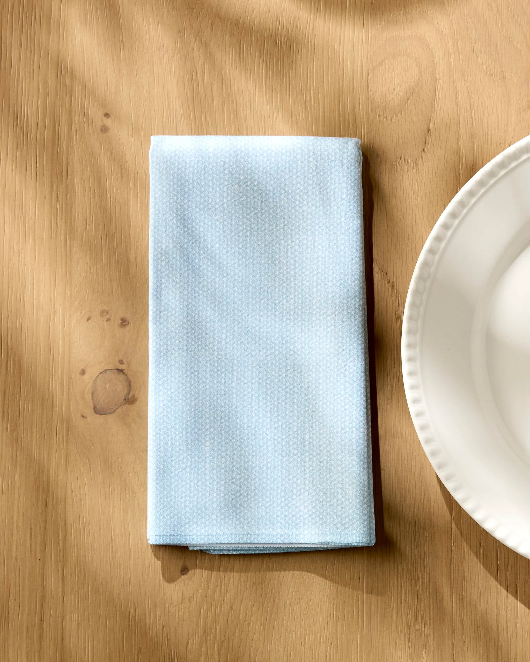 A Geometry Chambray Light Blue recycled dinner napkin with a subtle dotted pattern is folded on a wooden table beside a white ceramic plate.