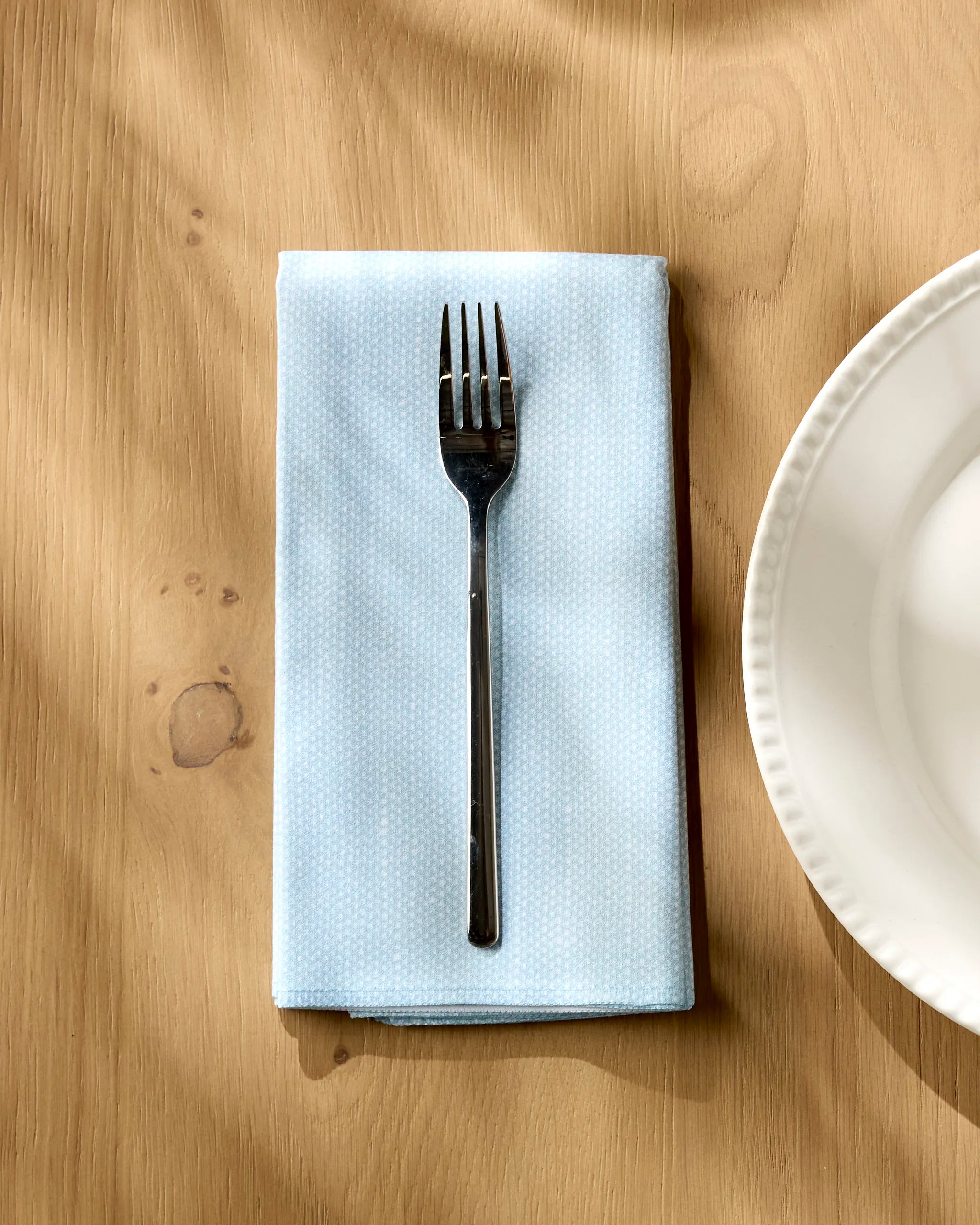 A metal fork rests on a folded Geometry Chambray Light Blue napkin, featuring a subtle dotted pattern, placed on a wooden table next to a white plate.