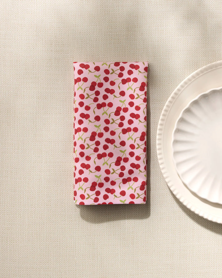 The GEOMETRY Cheery Cherries folded reusable dinner napkin, featuring a red cherry pattern on light pink, is displayed beside a white scalloped plate atop a textured cream tablecloth.