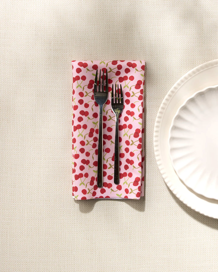 Two forks rest on a folded GEOMETRY Cheery Cherries reusable napkin, set beside two white scalloped plates atop a textured light-colored tablecloth.