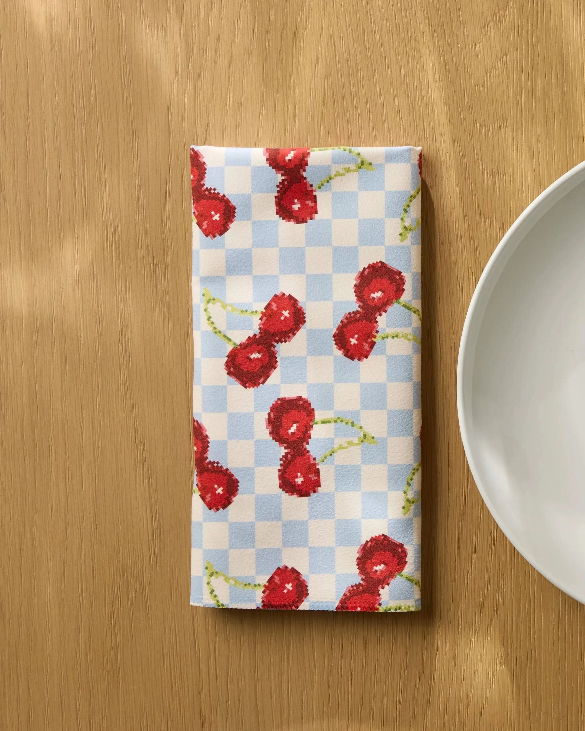 The Geometry "Cherry Cola Days" reusable table napkin, featuring a blue and white checkered pattern with red cherries, is folded and placed on a wooden table beside a white plate.