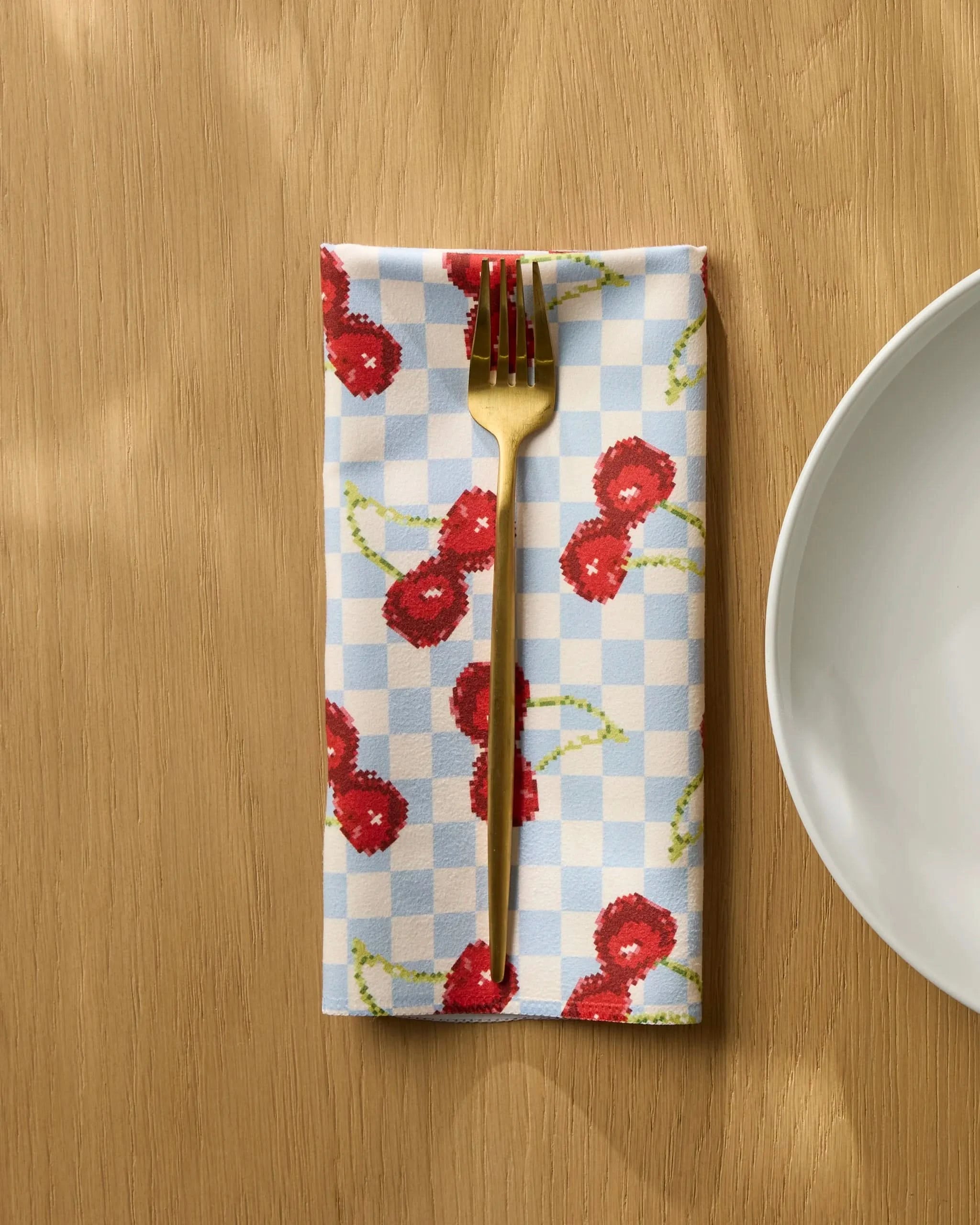 A gold fork rests on Geometry's Cherry Cola Days reusable napkin, featuring a red cherry pattern and blue checkered design, beside a white plate on a wooden table.