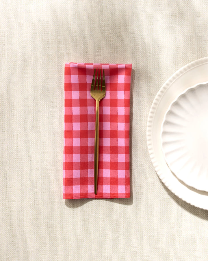 A gold fork rests on the GEOMETRY Cherry Gingham red and white checkered dinner napkin set, beside a white scalloped ceramic plate on a light textured tablecloth.