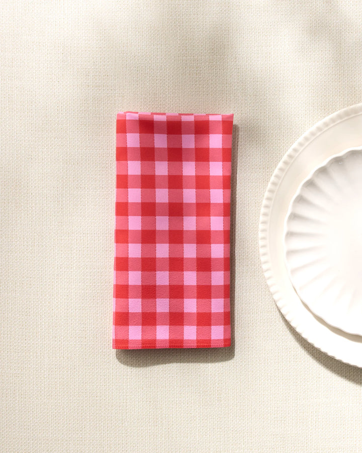 The Cherry Gingham napkin by GEOMETRY is neatly folded beside two stacked white scalloped plates on a light textured surface.