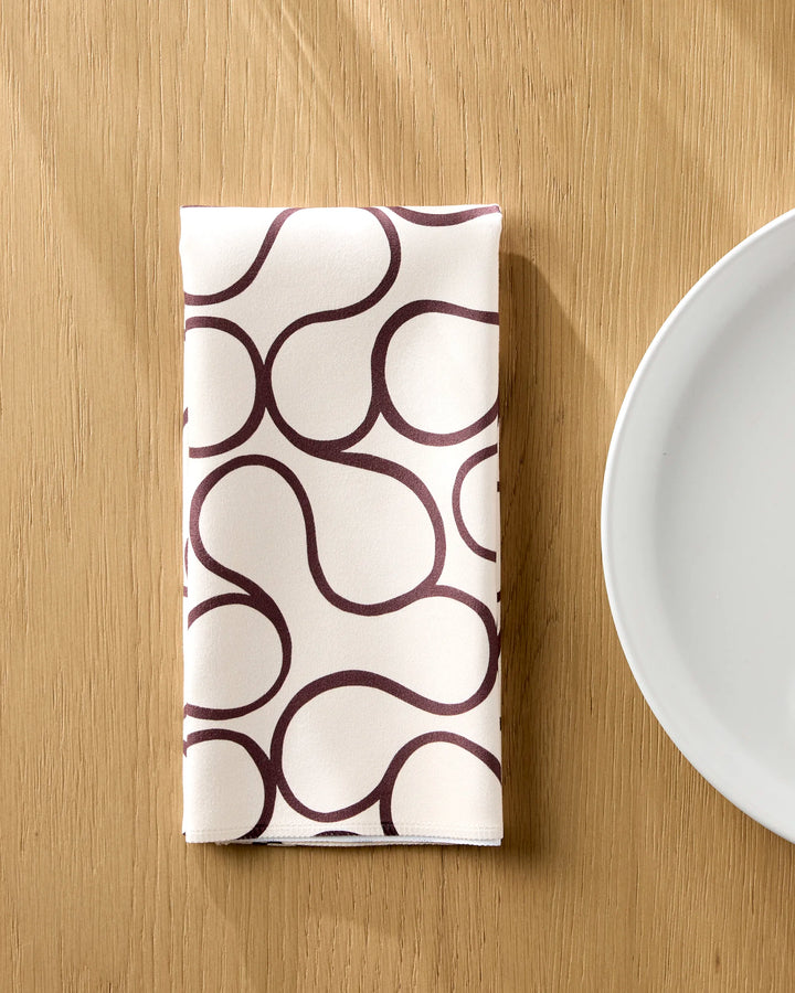 The Gerdadzy Coffee Swirl folded white reusable table napkin with a dark brown abstract pattern rests on a light wooden table beside a plain white plate.