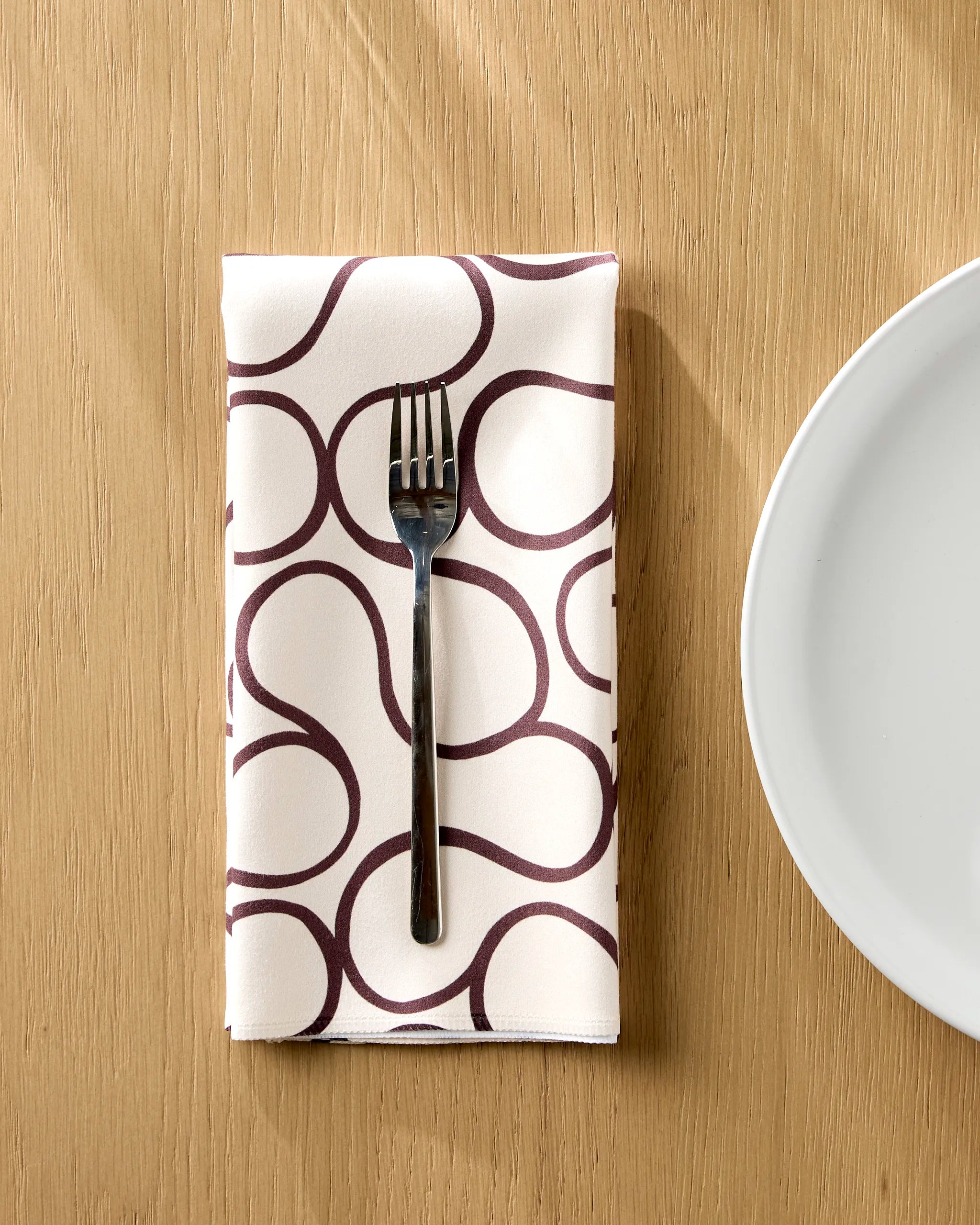 A metal fork rests on a folded Gerdadzy Coffee Swirl reusable napkin with a dark abstract pattern, set on a light wooden table beside a white plate.