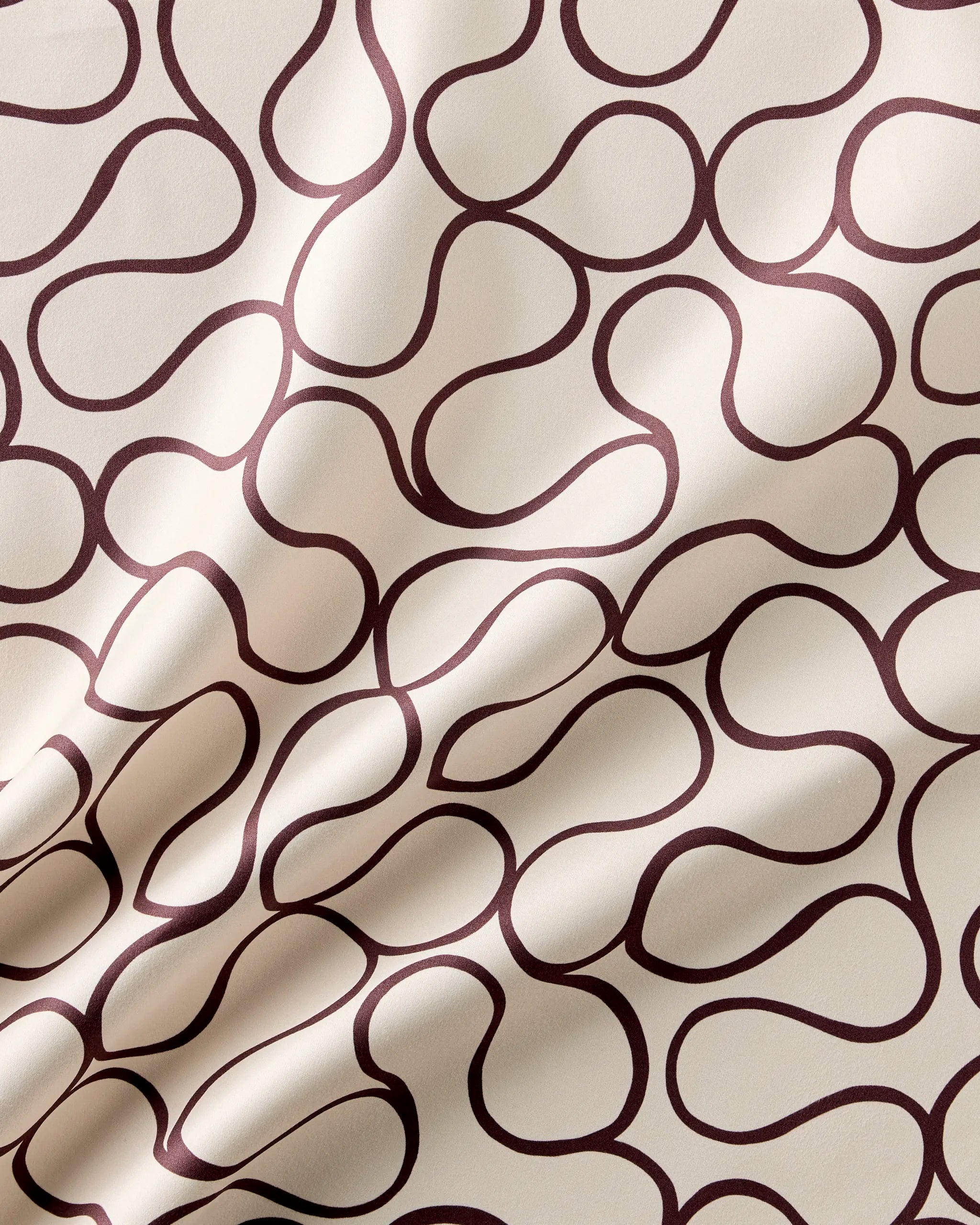 The Coffee Swirl reusable table napkin set by Gerdadzy features cream-colored fabric with a maroon swirling line design. Its slightly wrinkled texture adds depth, making this eco-friendly set both elegant and sustainable.