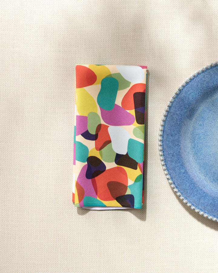 A GEOMETRY "Colorful Pebbles" recycled napkin, featuring an abstract pattern, is neatly folded beside a textured blue plate on a light woven tablecloth.