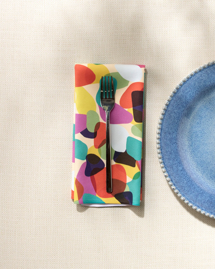 A silver fork sits on GEOMETRY’s Colorful Pebbles recycled napkin, placed beside a blue scalloped-edge plate on a light textured surface.