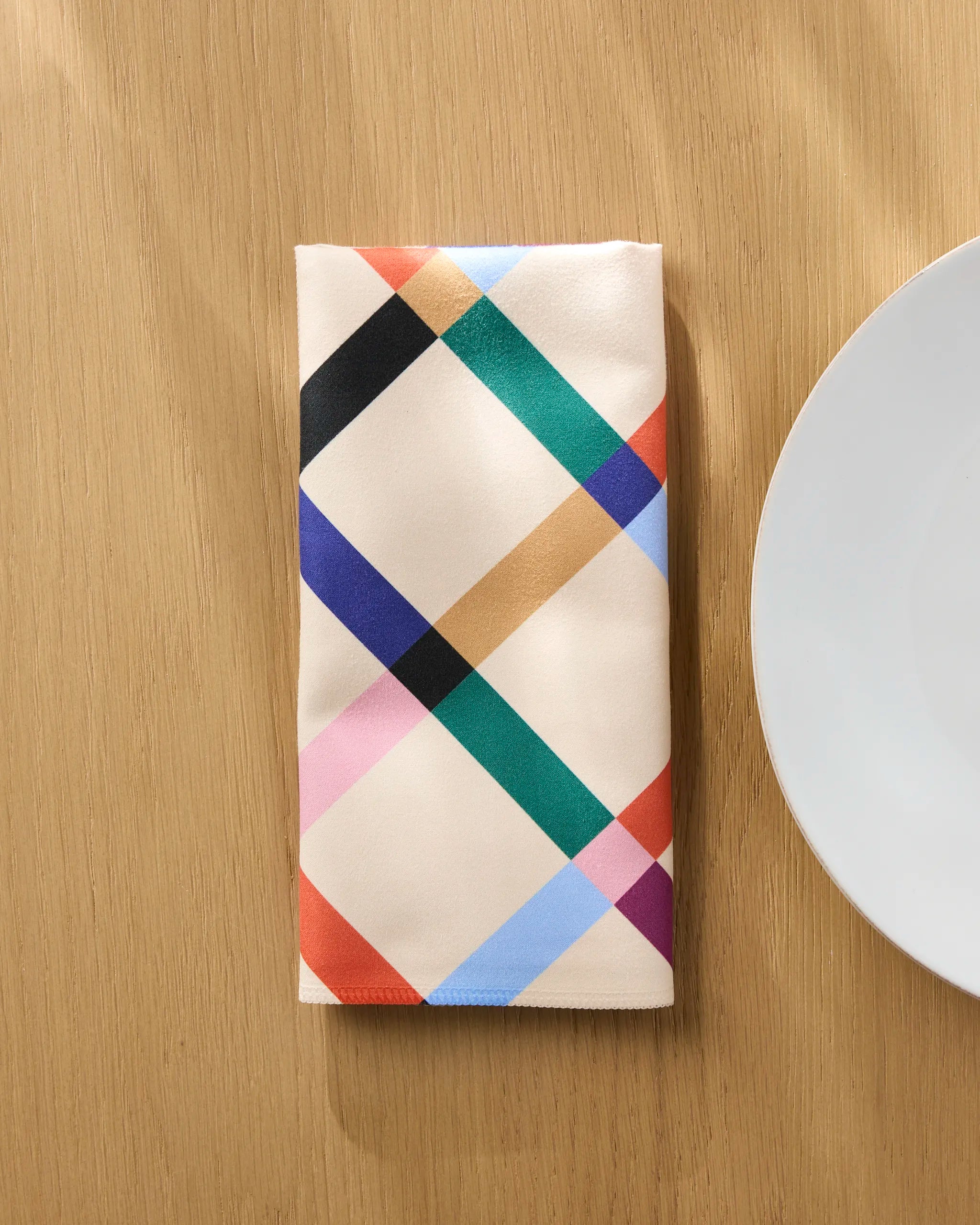 The Cool Grid reusable cloth napkin by Jennifer Bouron, featuring a colorful geometric pattern, is neatly folded on a light wooden table beside a white plate.