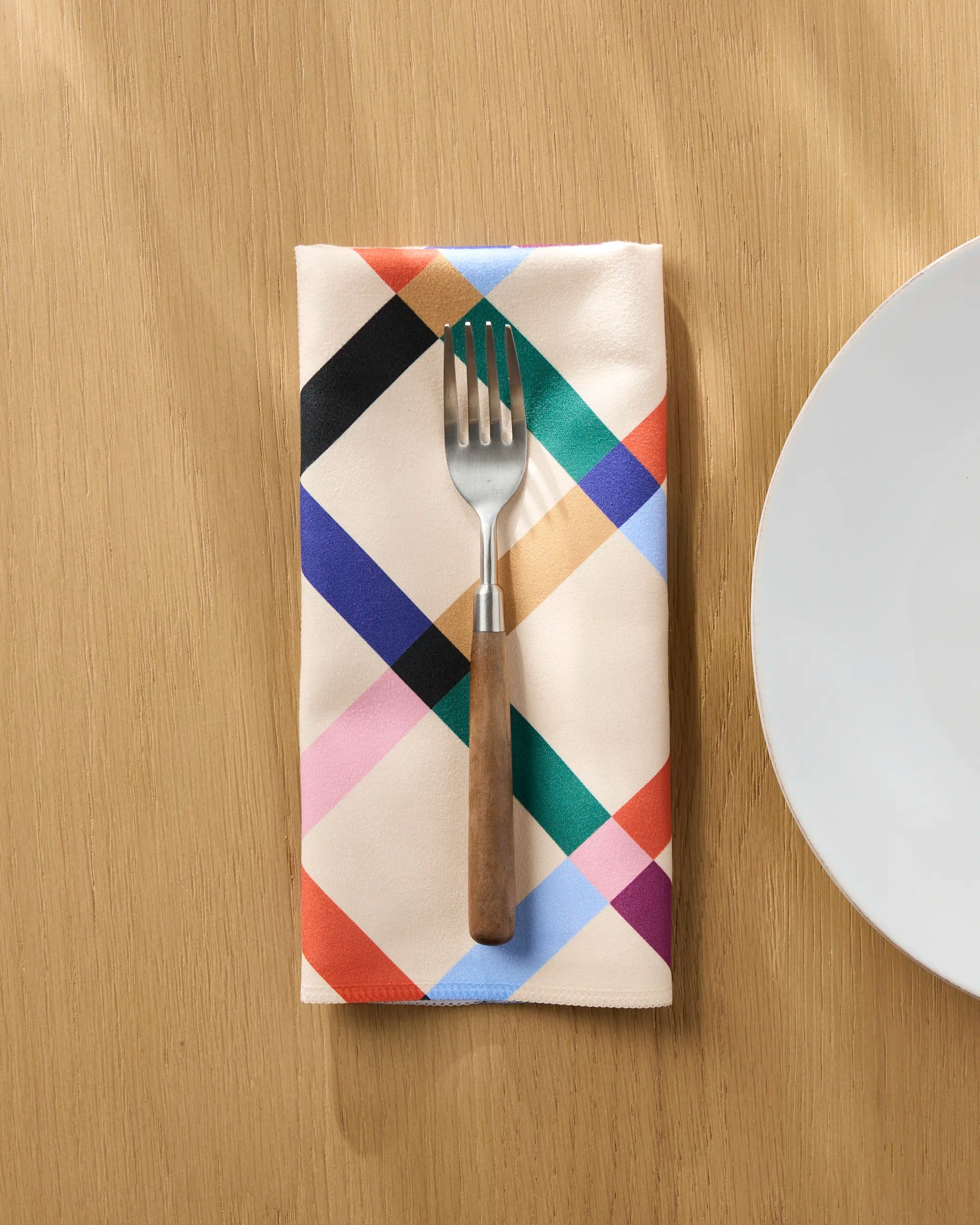 The Jennifer Bouron Cool Grid reusable cloth napkin with a colorful plaid pattern is folded on a light wooden table, holding a fork with a wooden handle beside a white plate.