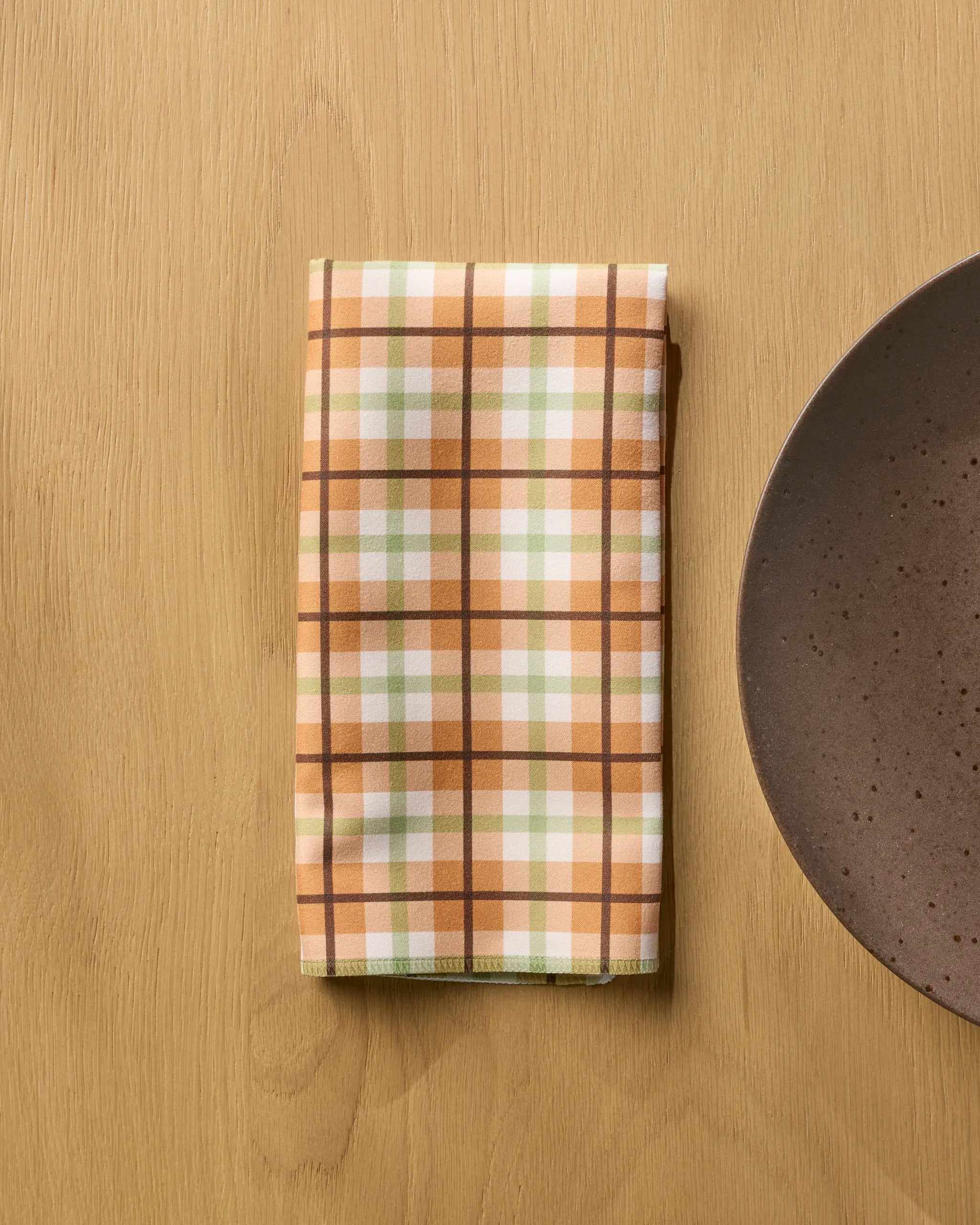 A folded plaid napkin from the Geometry Country Kitchen set, with brown, beige, white, and light green stripes, rests on a light wooden table beside a round brown ceramic plate.