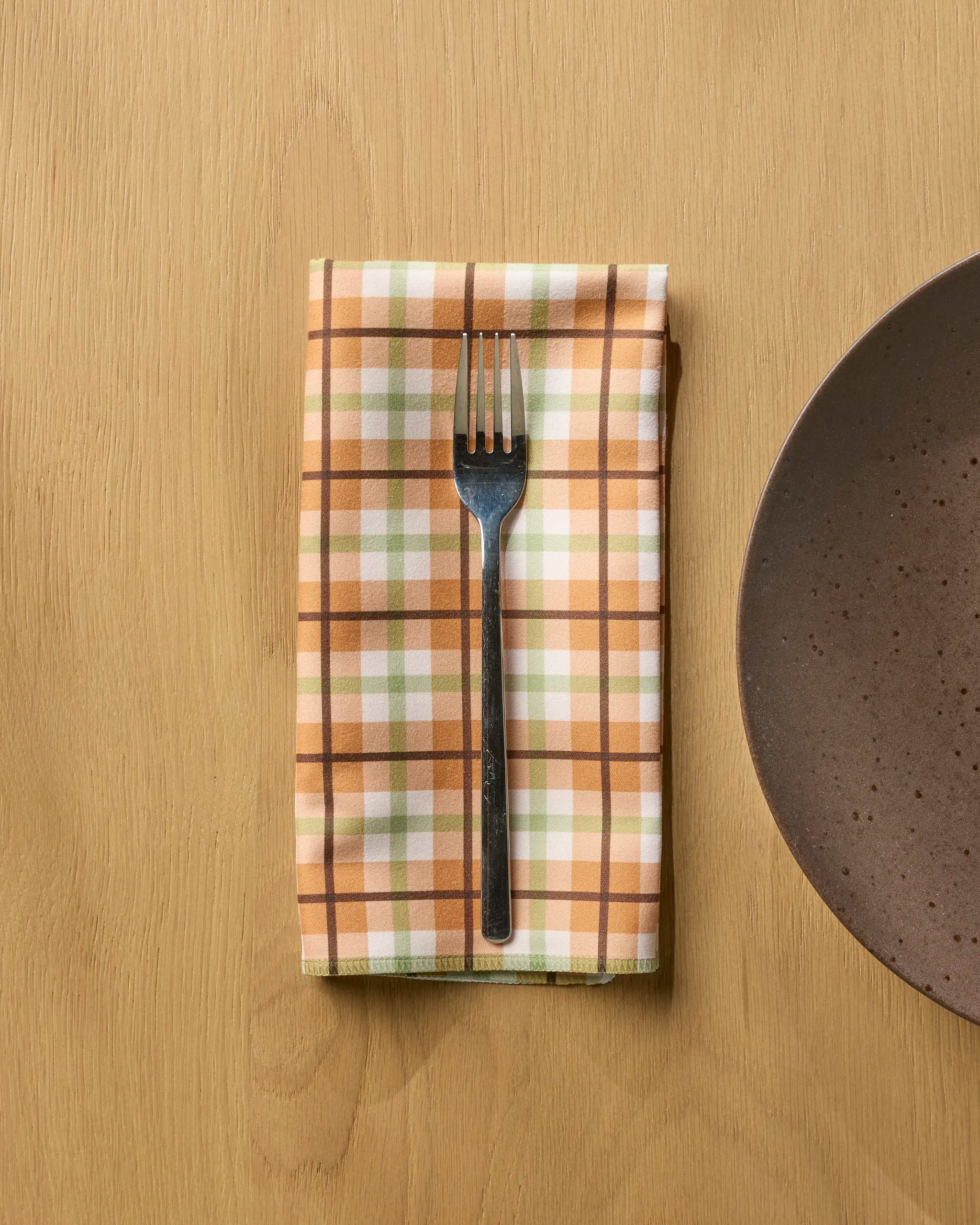 A metal fork rests on a folded Geometry Country Kitchen orange and white plaid reusable napkin, placed on a wooden table beside a brown ceramic plate.